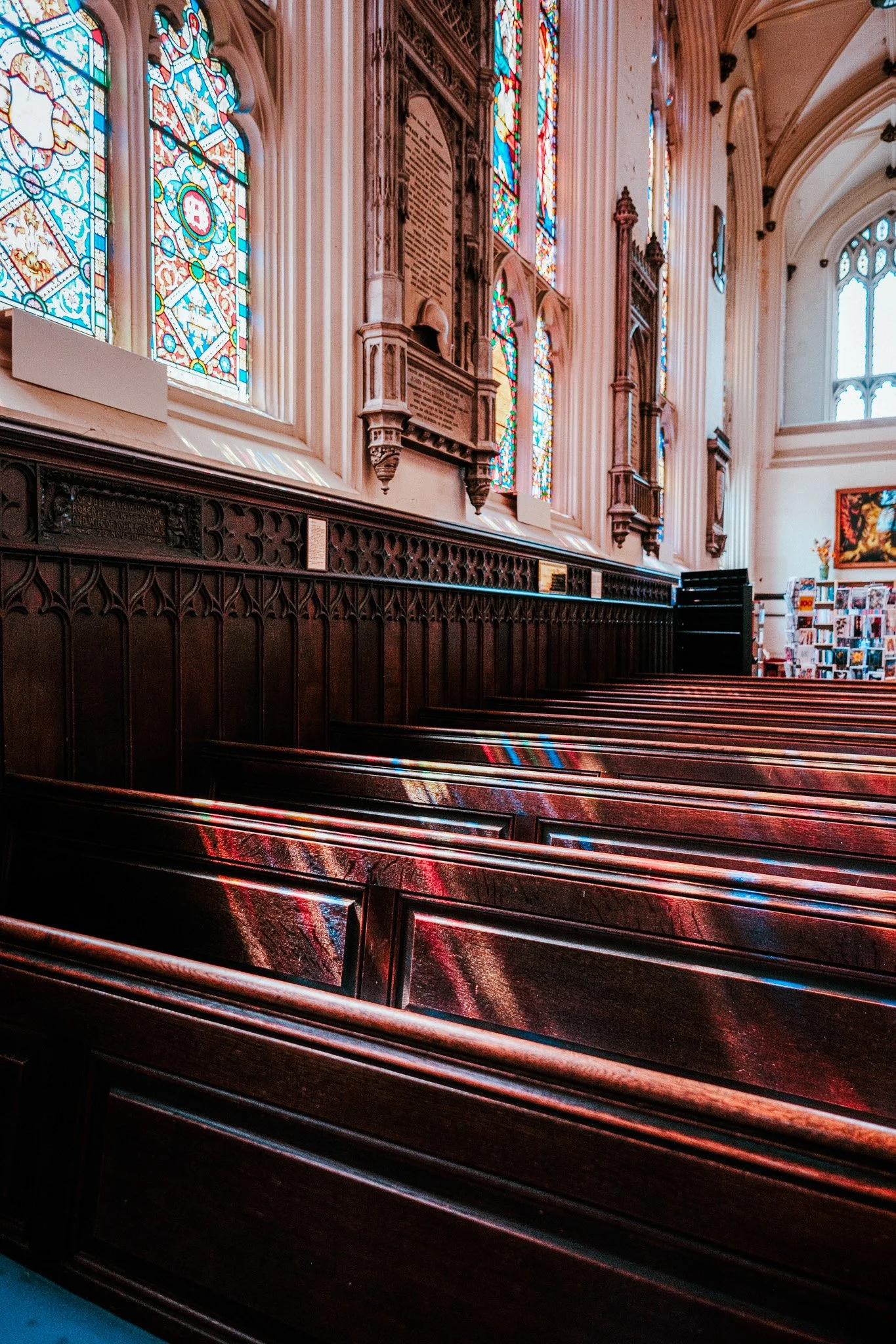 Interior of a church with wooden pews, stained glass windows, intricate woodwork, and a bookshelf with books and magazines.
