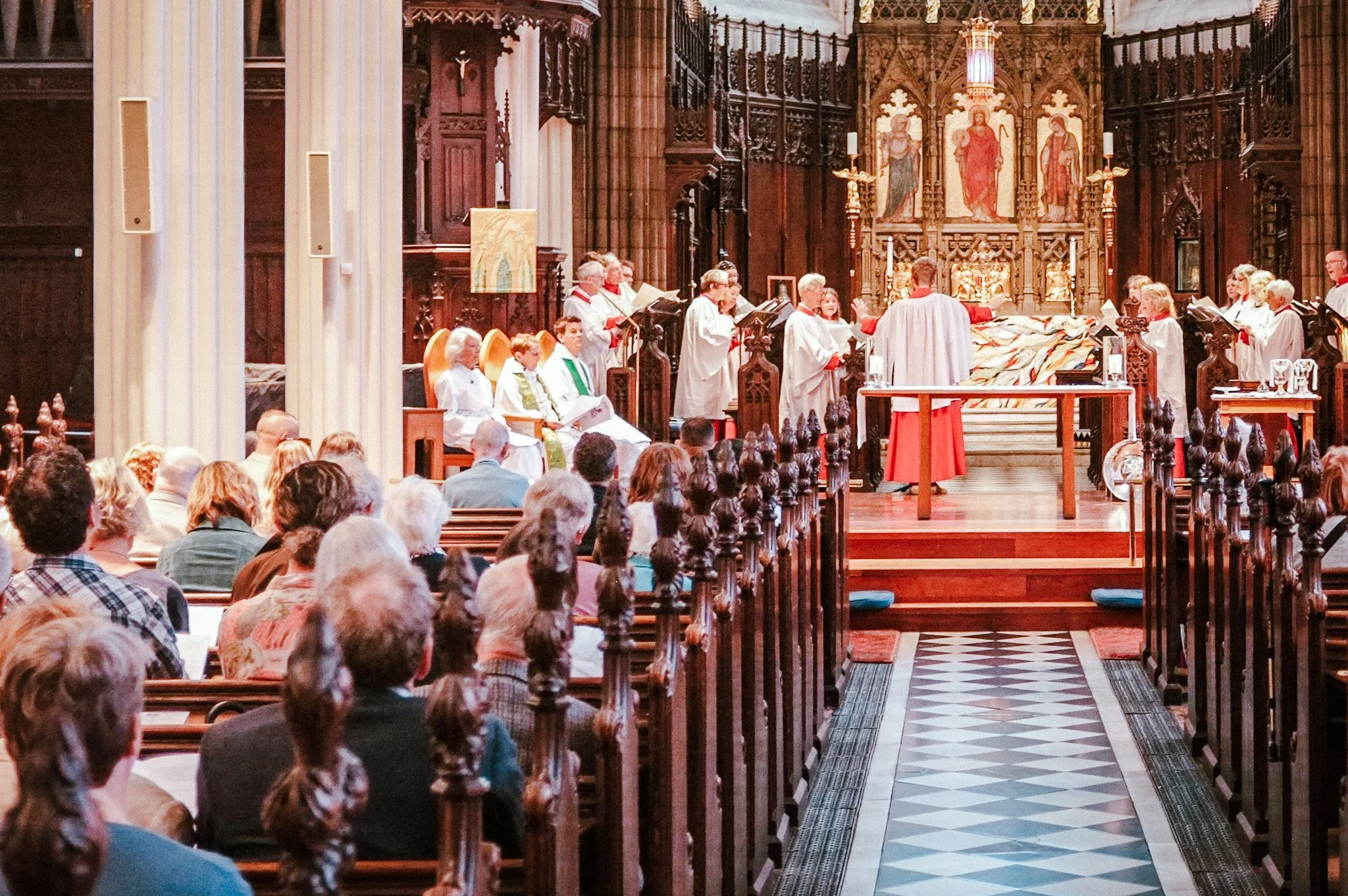 Church service with choir singing at altar, congregation seated in pews, ornate woodwork and religious artwork on wall.