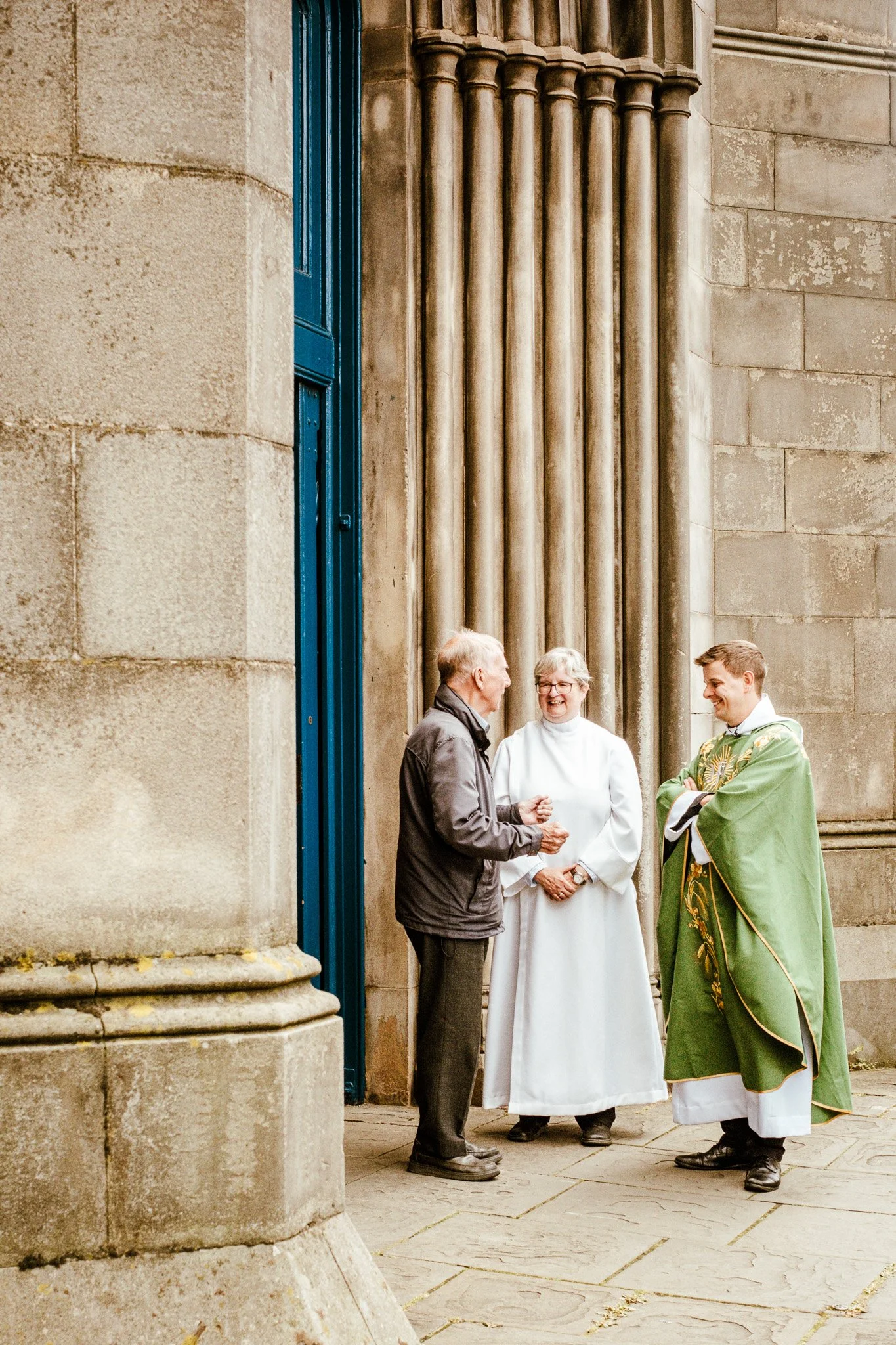 Four people are standing outside a stone building having a conversation. One is a man in a gray jacket, an older woman in a white religious habit, and a young man in traditional robes with religious symbols.