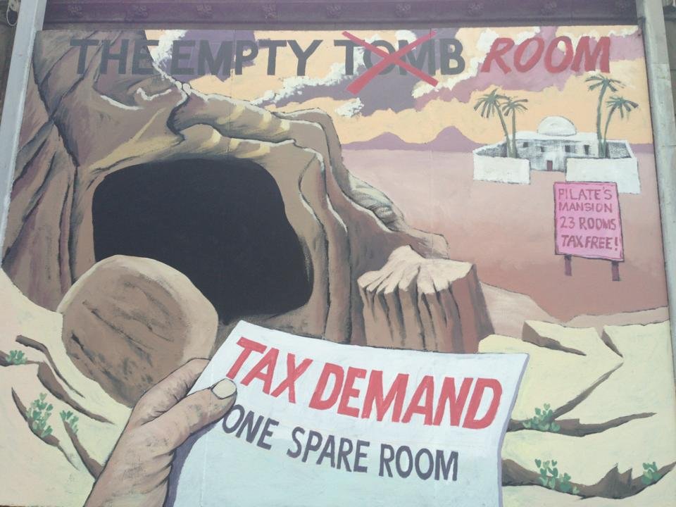 A mural showing a large limestone cave with a sign advertising 'Pilate's Mansion, 23 rooms, tax-free.' The mural includes an image of a hand holding a sign that reads 'Tax Demand, One Spare Room.' The top part of the mural has 