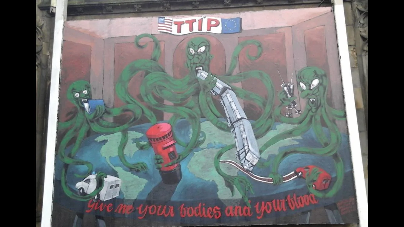 Street art mural featuring green tentacled monsters with angry expressions, holding various rescue tools like a fire hose, walkie-talkie, medical syringe, and rescue equipment. The mural includes the text 'TTIP' with flags of the United States and Eu