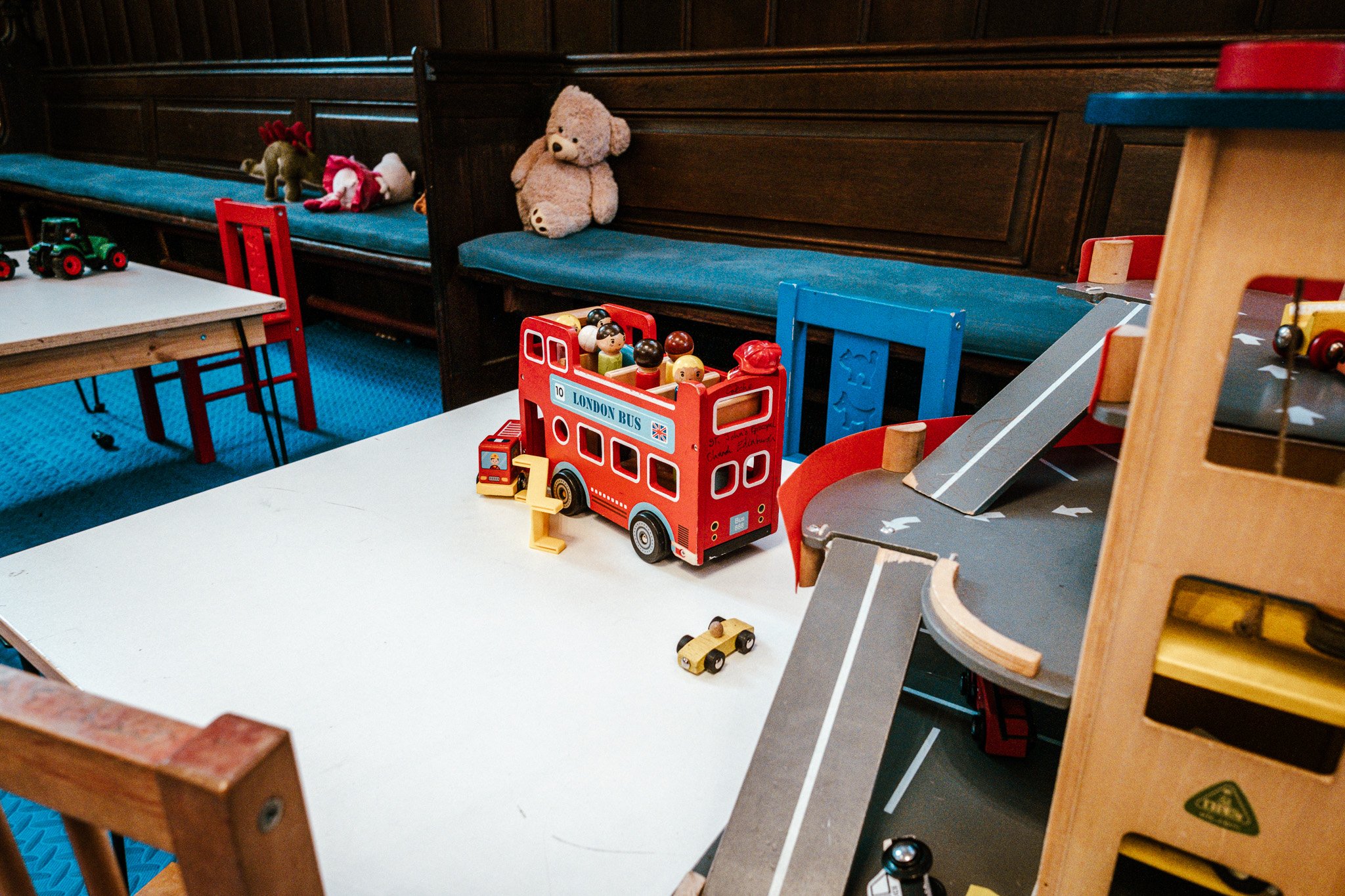 Children's play area with wooden toys, green toy tractors, plush teddy bear, and toy bus on tables and shelves with toys in a room with blue and red chairs
