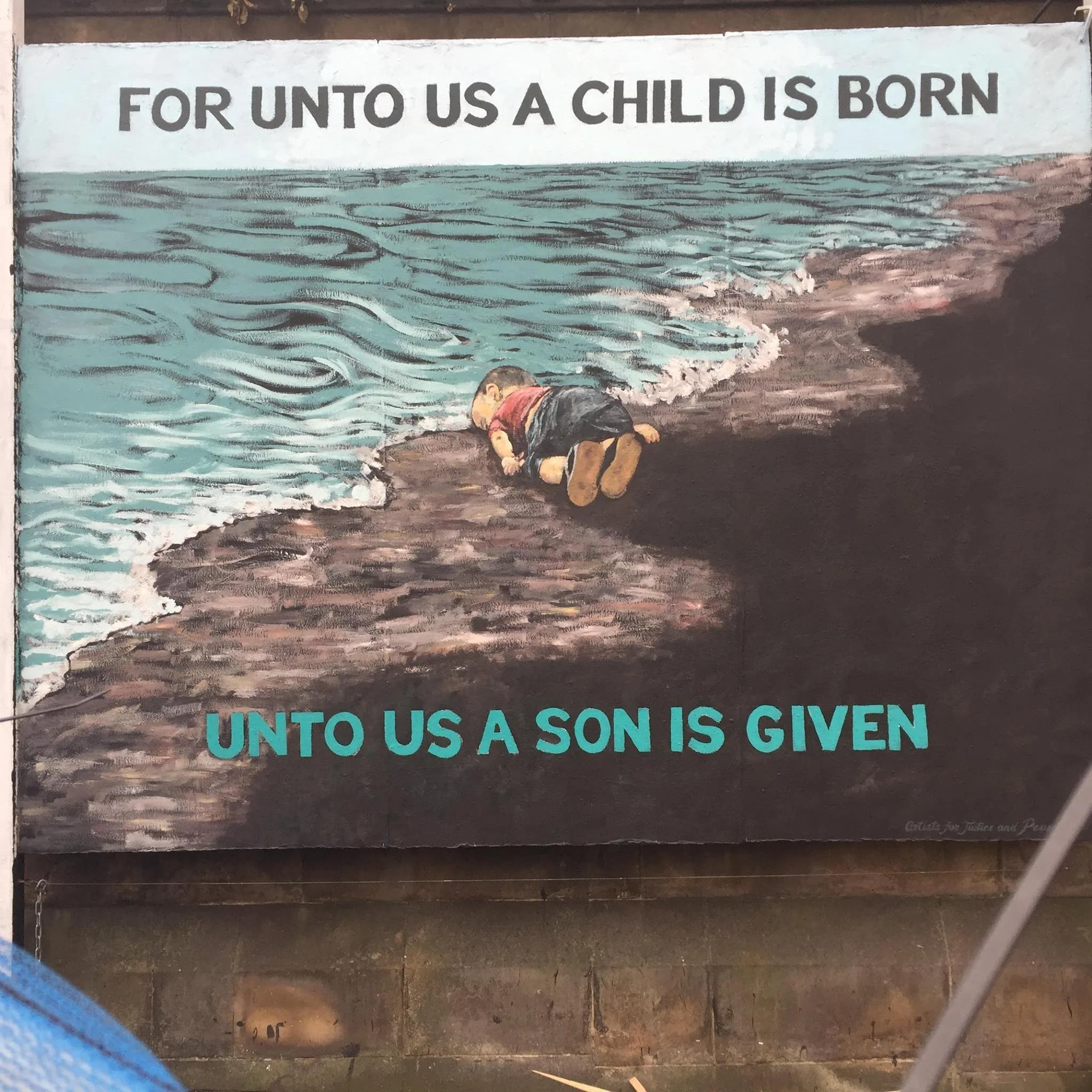 A mural with the text "For unto us a child is born" at the top, depicting a boy wearing a red shirt and black shorts, kneeling on the ground by the water’s edge, with waves and shoreline visible.
