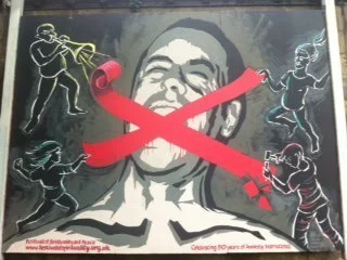 Street art mural depicting a person with a large central face and a red crossed-out banner across it, with figures of children playing or fighting on either side.