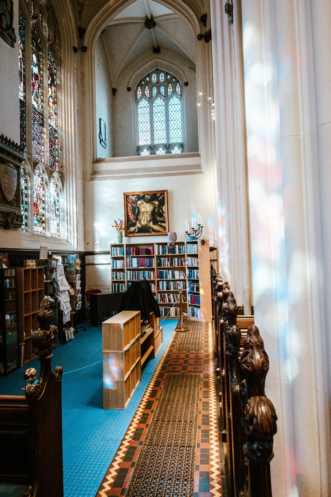 Interior of a church with stained glass windows, a bookshelf, artwork on the walls, and wooden pews along a patterned tile and carpeted aisle.