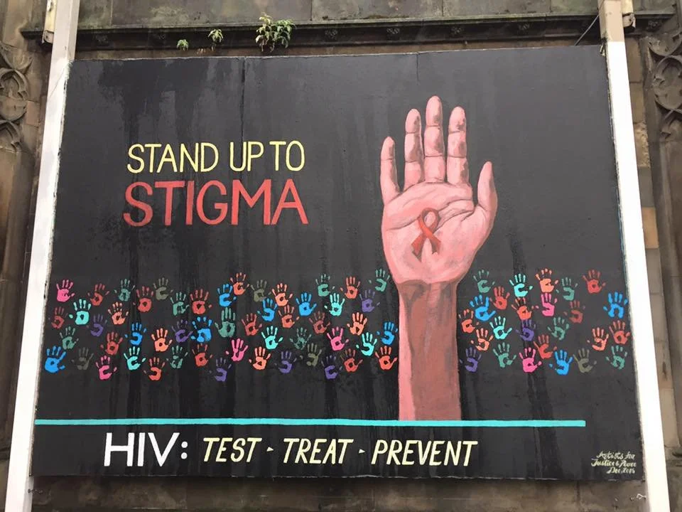 A sign with the message 'Stand Up to Stigma' and a large outstretched hand with a ribbon painted inside the palm. The sign features multiple colorful handprints and the message 'HIV: Test - Treat - Prevent' in bold letters.