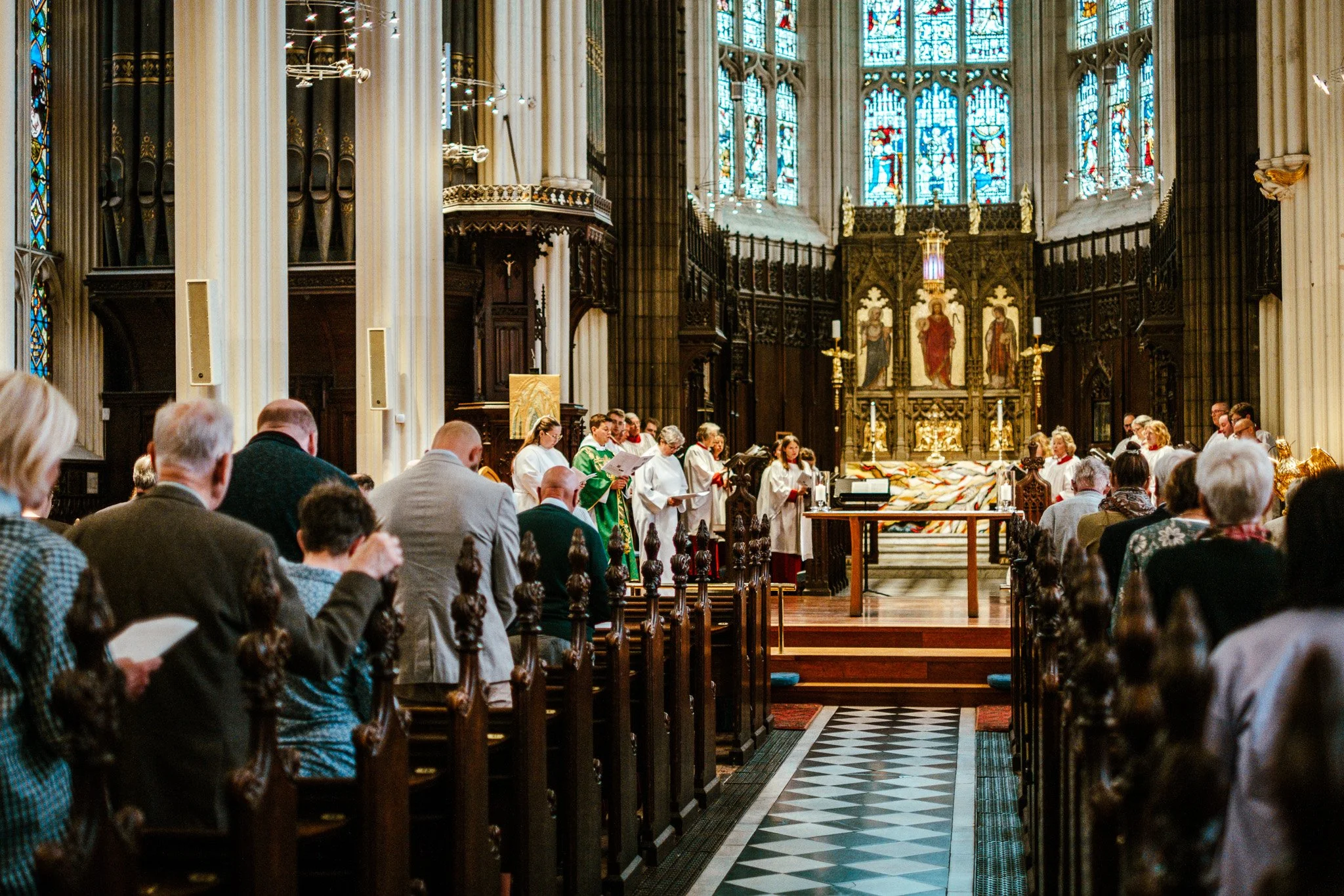 Inside a church during a religious service, with congregation members seated, altar at the front, stained glass windows, and clergy dressed in green robes.