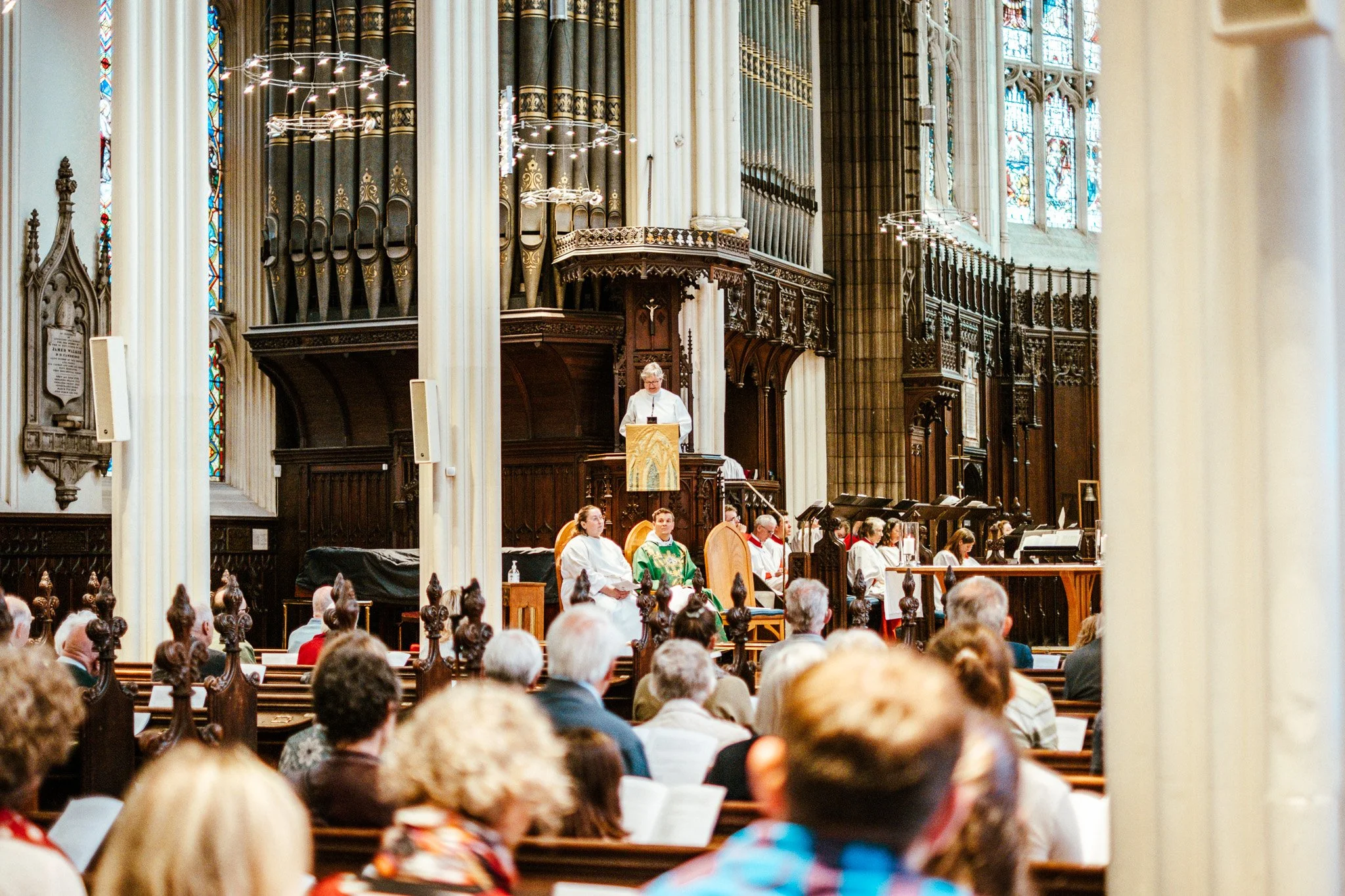 Inside a large church or cathedral during a service, with a priest or minister standing at the pulpit, congregation members seated in pews, and stained glass windows and ornate woodwork in the background.