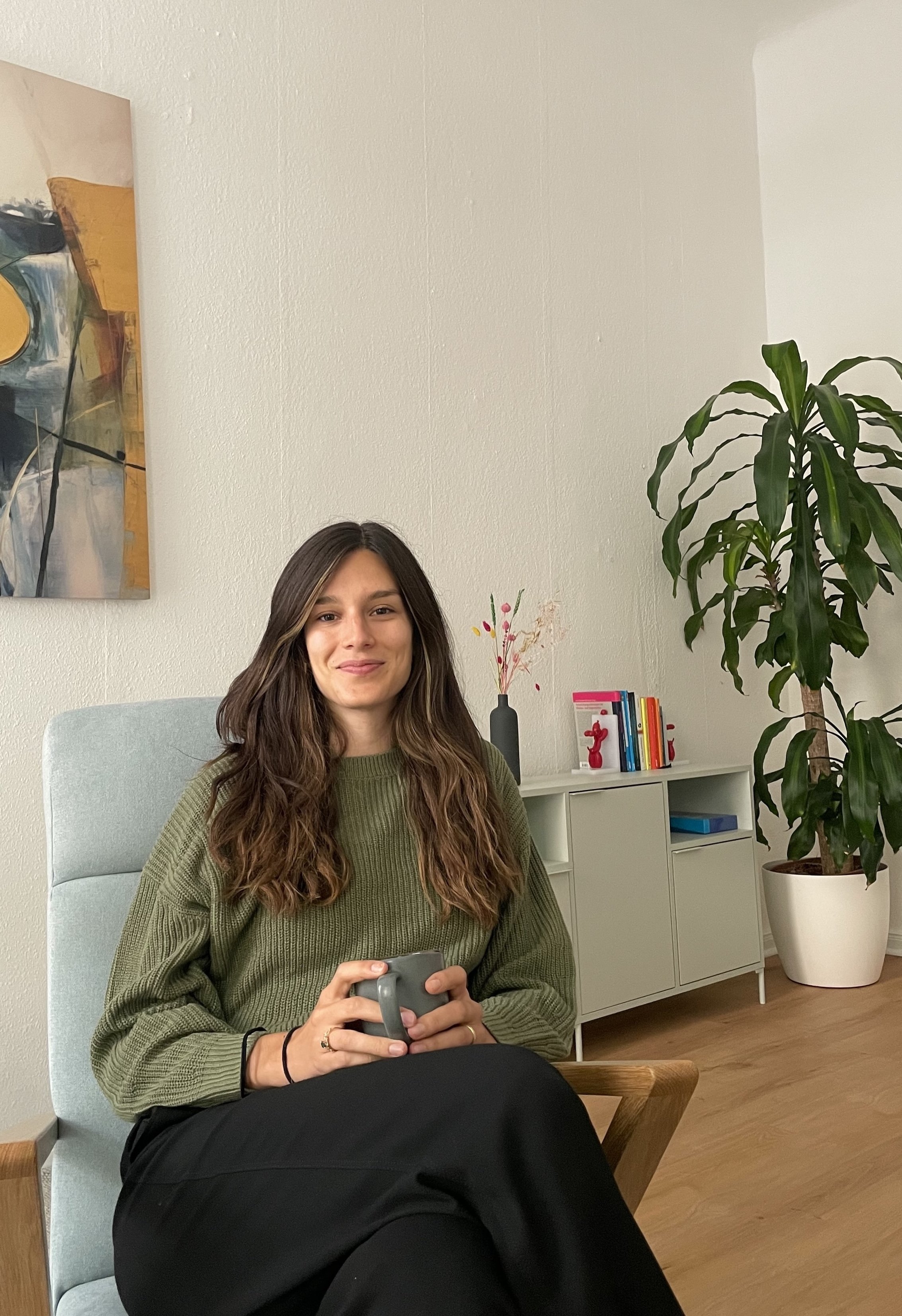 Die Therapeutin (eine junge Frau mit braunen Haaren) sitzt in einem Therapierum, hält eine graue Tasse und lächelt. Im Hintergrund sind eine hellgrüne Kommode mit bunten Büchern, eine große Topfpflanze und eine abstrakte Wandkunst sichtbar.