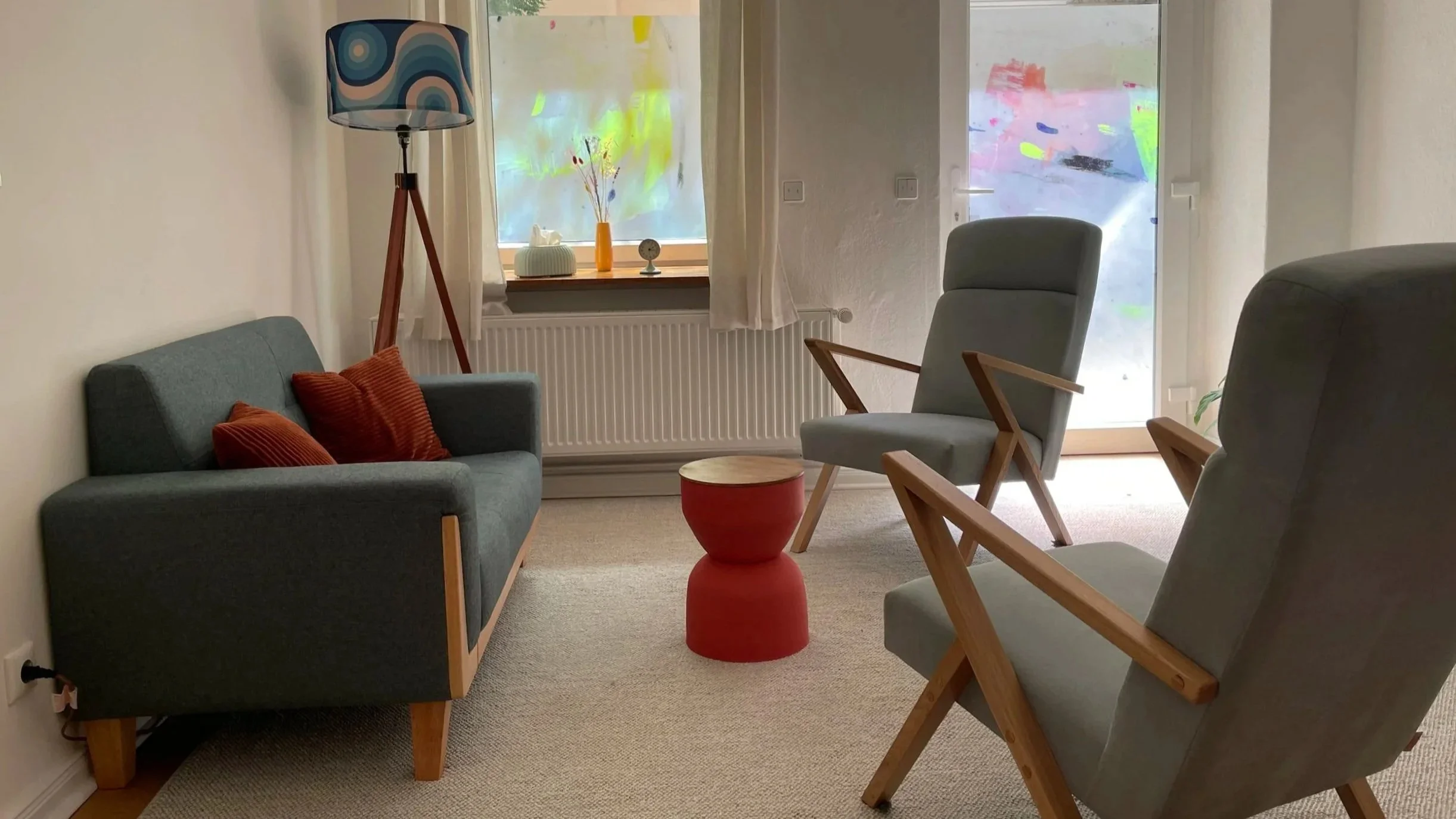 Gemütliches Wohnzimmer mit grauen Möbeln, roten Kissen, einem roten Beistelltisch, einem Stehlampen mit buntem Lampenschirm, und Fenstern mit weißen Vorhängen, die auf eine bunte, abstrakte Tür oder Wand hinter Glas schauen.