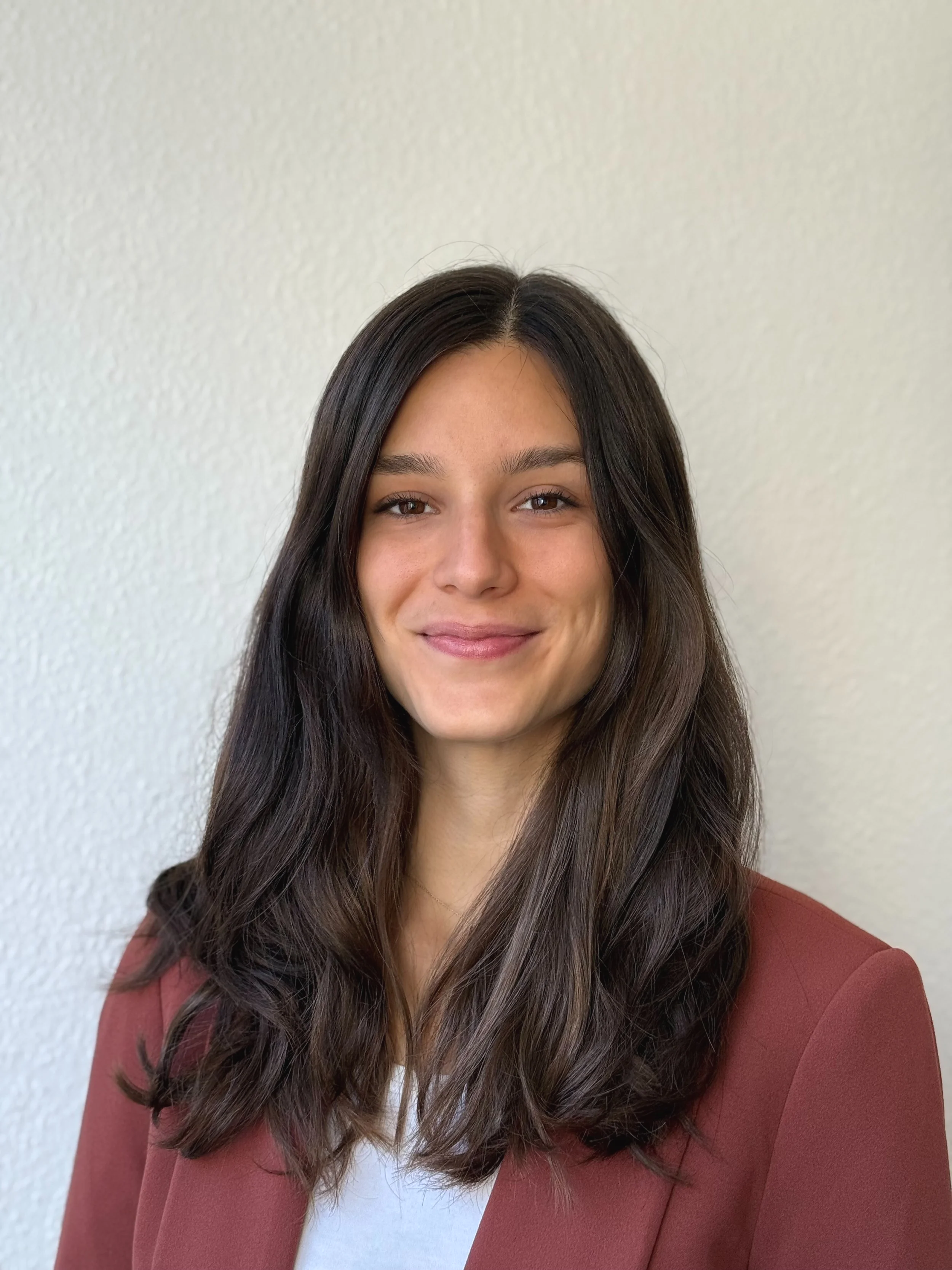 Nahaufnahme einer lächelnden jungen Frau mit langen braunen Haaren, dunklen Augen, rotem Blazer und weißem Shirt vor einer hellen Wand.
