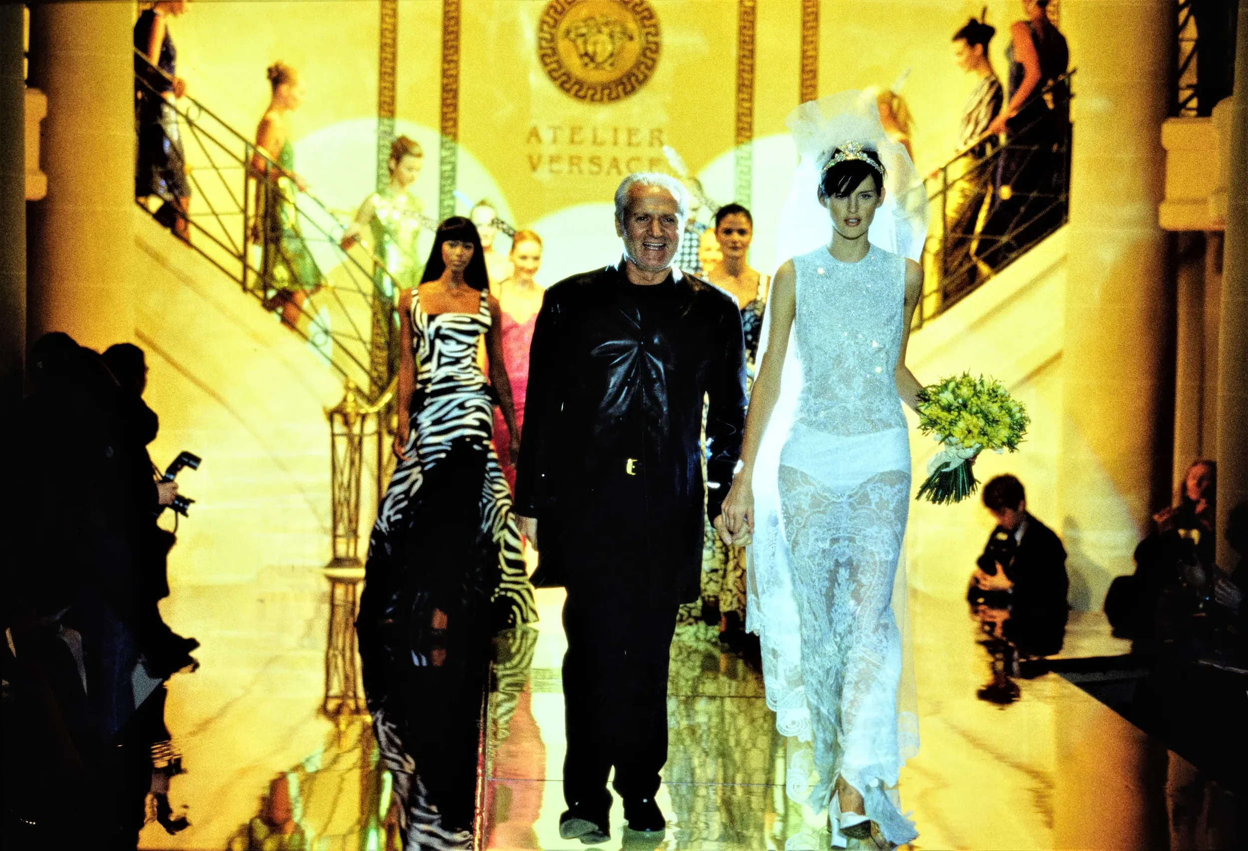 People walking down a runway with models wearing fashionable clothing, including a bride with a bouquet, at a show in a venue with a sign reading 'Atelier Versace' in the background. *Versace Atelier 1996