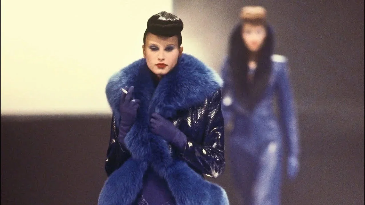 Fashion-forward woman with dark hair in a sleek updo, wearing a luxurious blue fur-collared coat, purple gloves, and bold makeup, with another woman dressed in a similar style blurred in the background. *Givenchy 1998