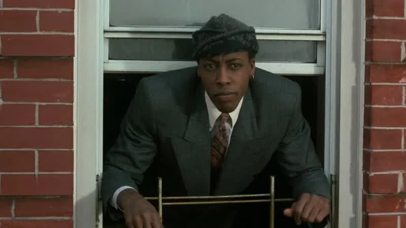 Man in a suit and tie leaning out of a window with a brick wall on either side.