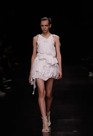 Model walking on runway in a white ruffled dress and beige high-heeled sandals. *Ann Demeulemeester 2009