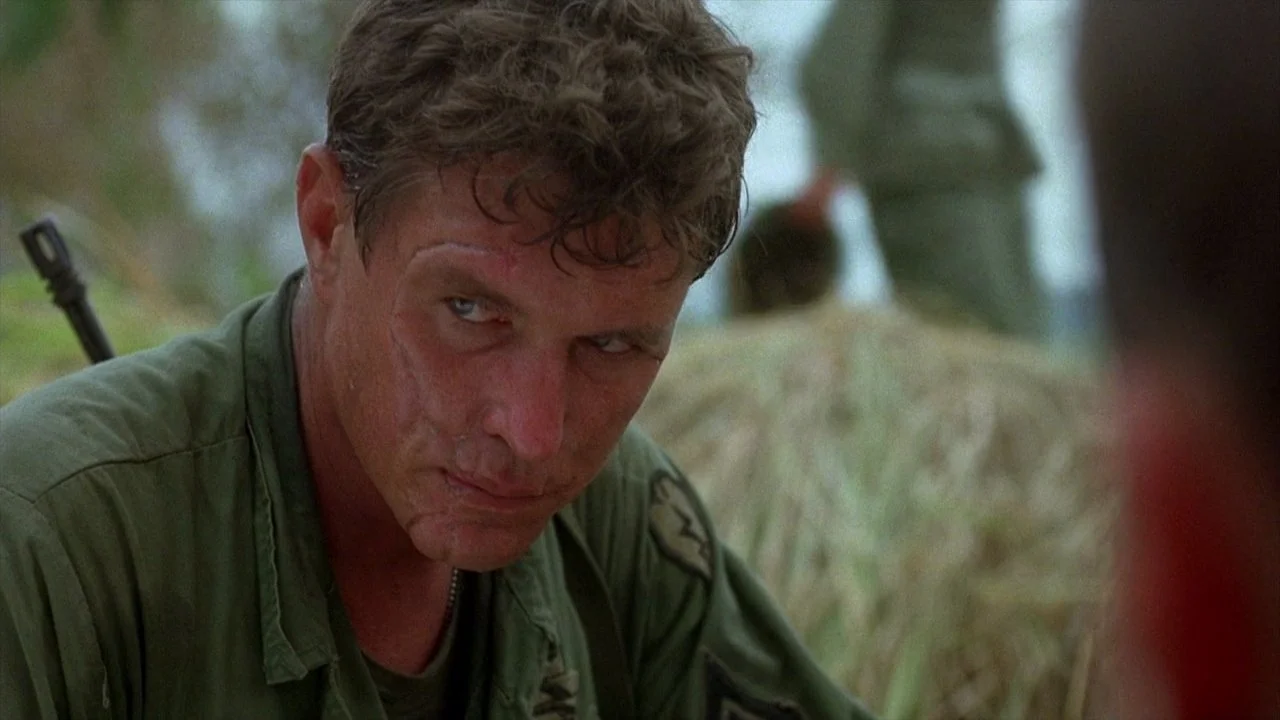 A man in military uniform looking intense, with dirt on his face and a rugged background.