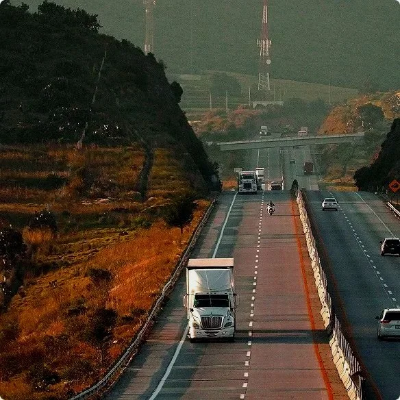 View of a divided highway with vehicles, including a semi-truck, cars, and a motorcycle, passing through a hilly area with trees and a bridge visible in the background.