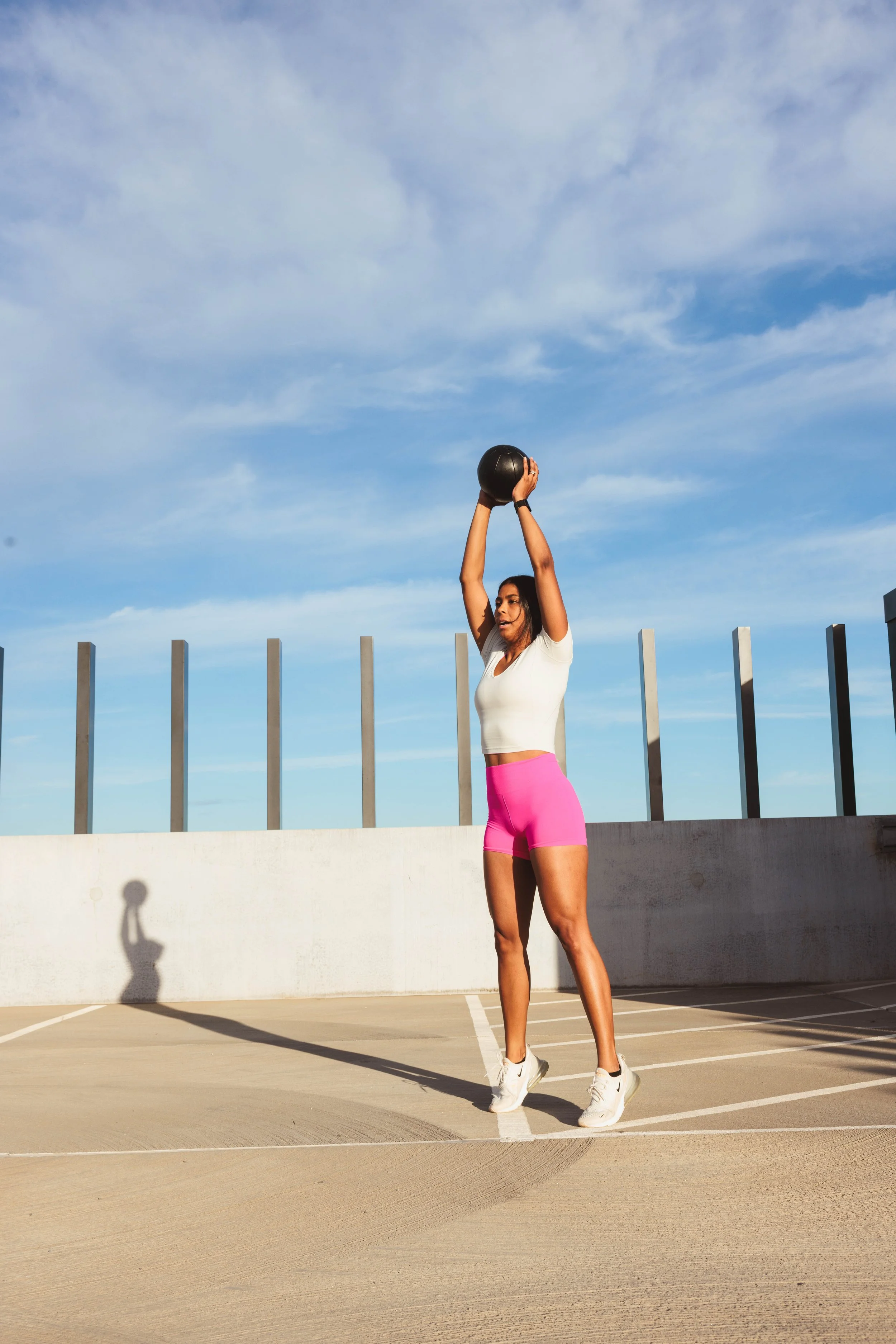 A woman wearing a white t-shirt and pink shorts is outdoors on a sport court, lifting a black medicine ball above her head while her shadow is cast on the wall behind her against a blue sky with clouds.