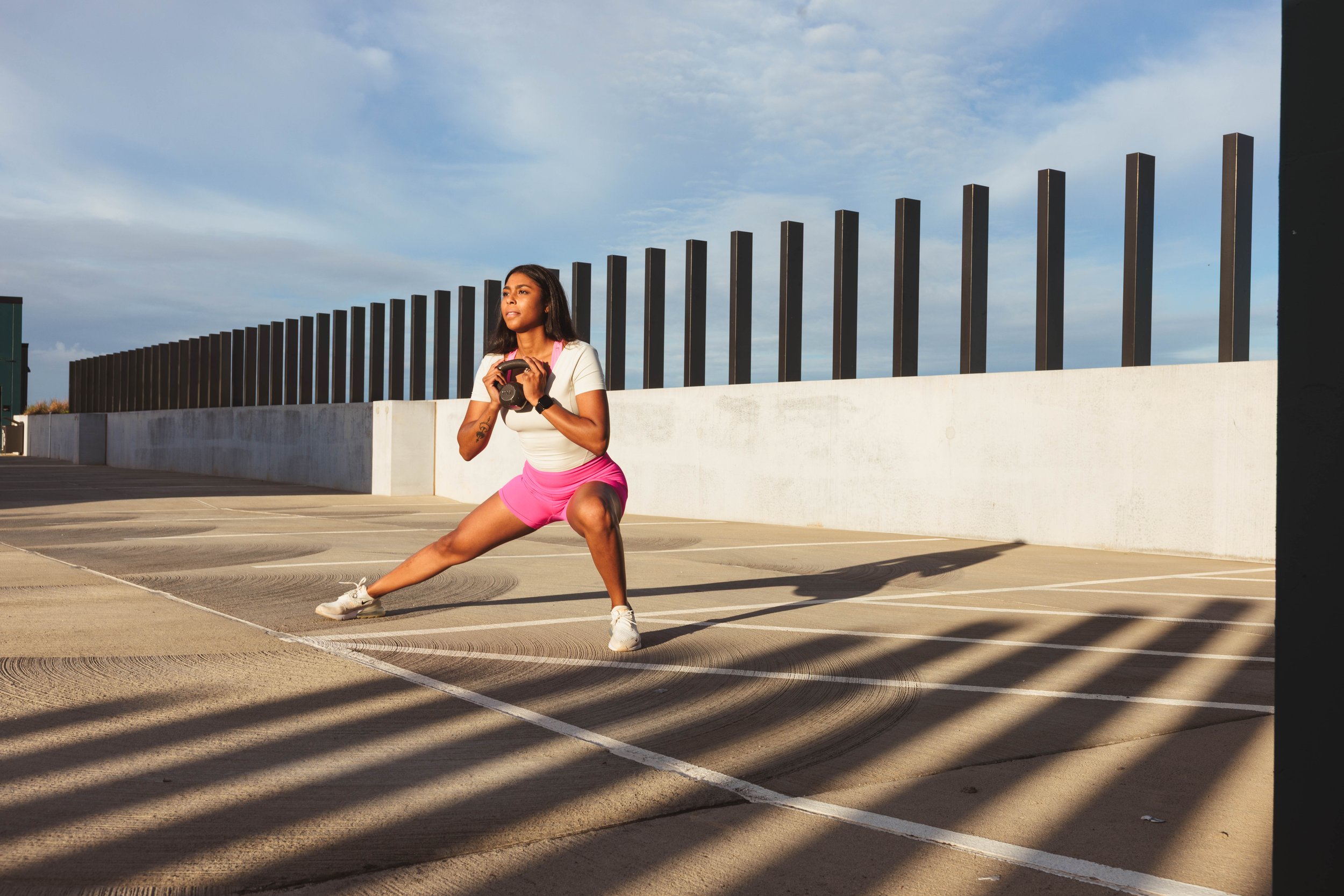 A woman in pink shorts and a white top is exercising on an empty parking lot during daytime, holding a kettlebell close to her chest while lunging forward with her left leg. There is a tall black fence and a blue sky with scattered clouds in the background.