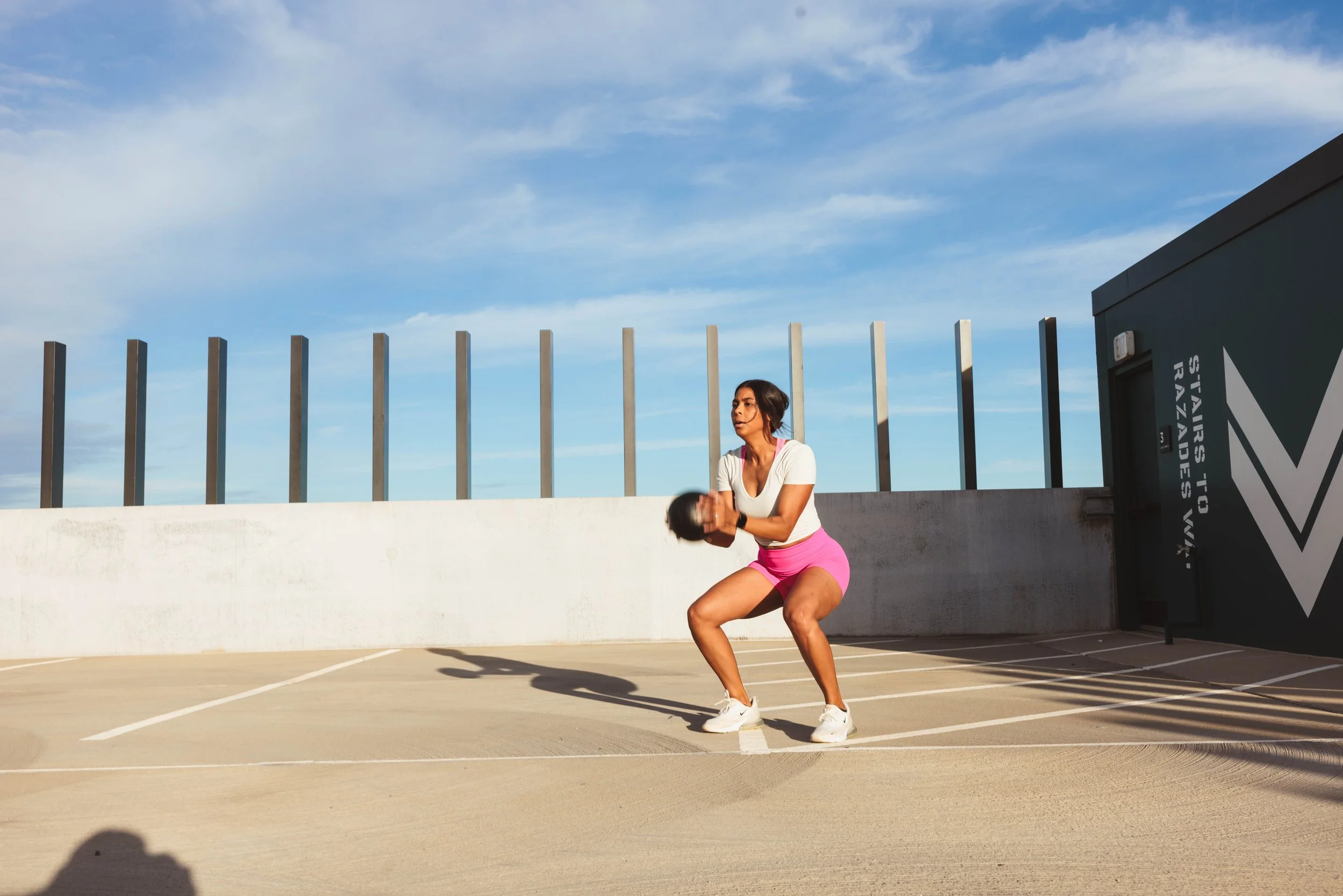 A woman in athletic clothing performing lunges in an outdoor parking lot near a tall fence and a building with a sign.