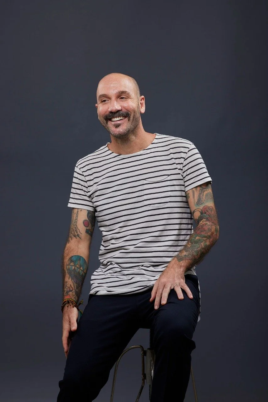 Vinicius Malinoski photo. He is a smiling, tattooed man with a mustache wearing a striped t-shirt sitting on a stool against a dark gray background.