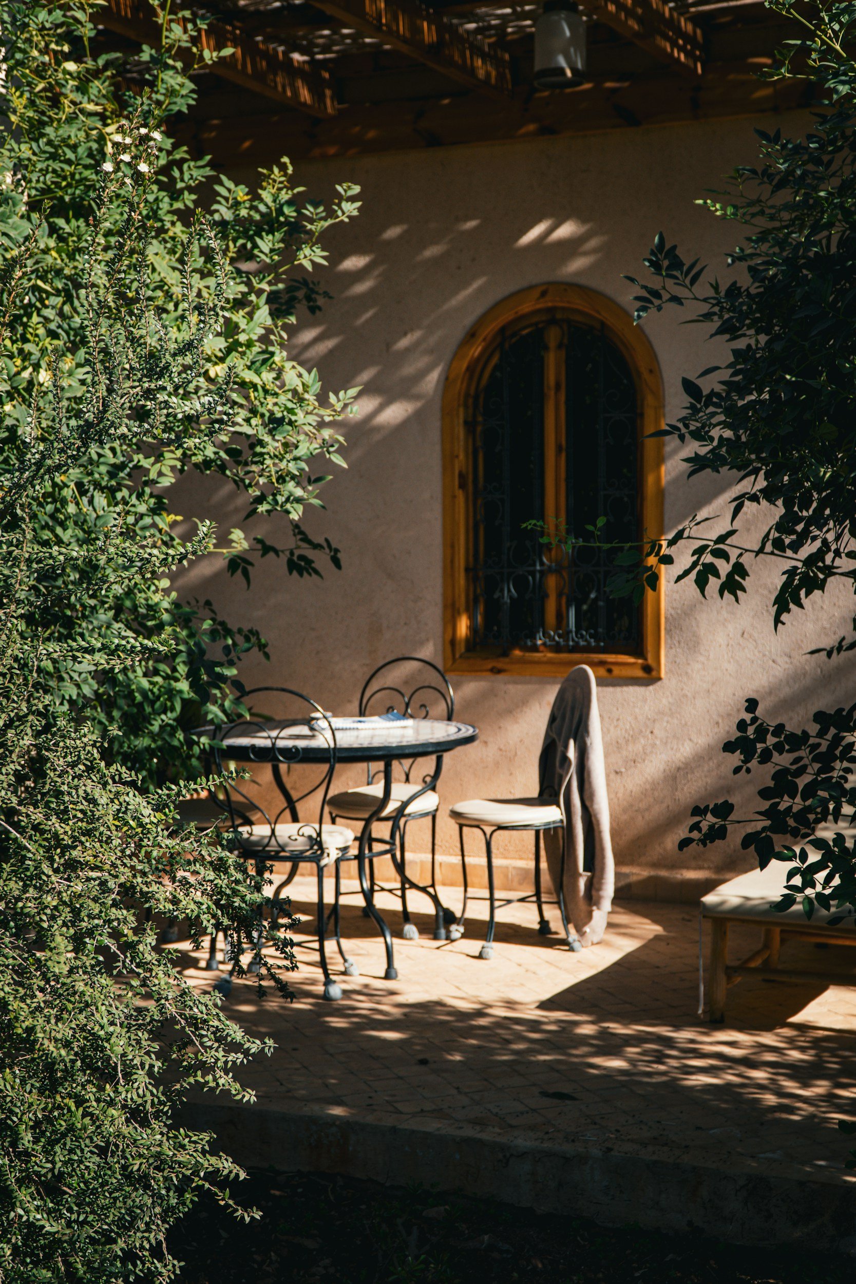 Outdoor patio with a circular metal table and four chairs, surrounded by green bushes and shaded by overhead tree branches, with a window on a stucco wall in the background.