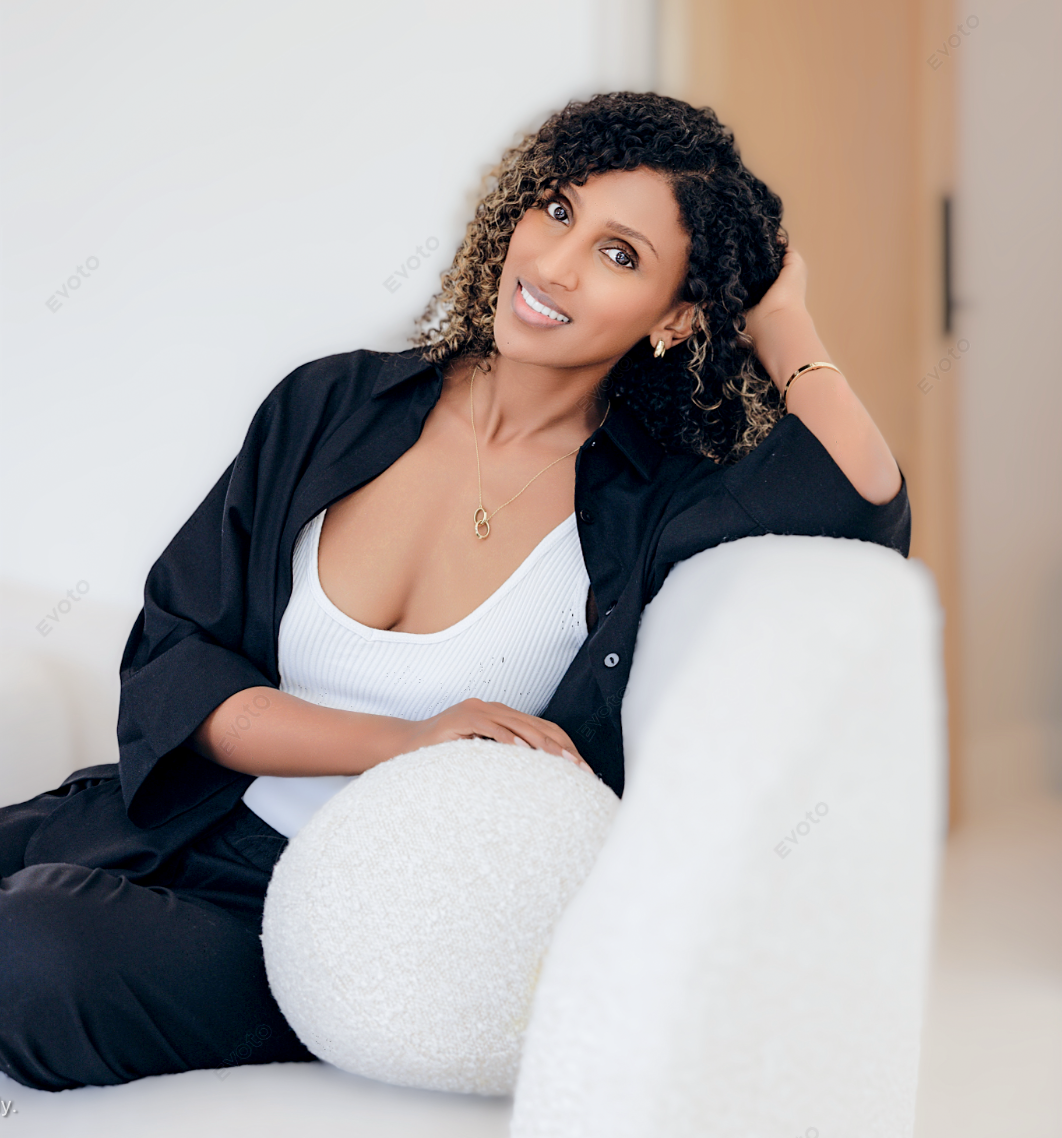 Woman with curly hair sitting on a white couch, resting her arm on a textured pillow, wearing a black shirt over a white top, smiling and looking at the camera.