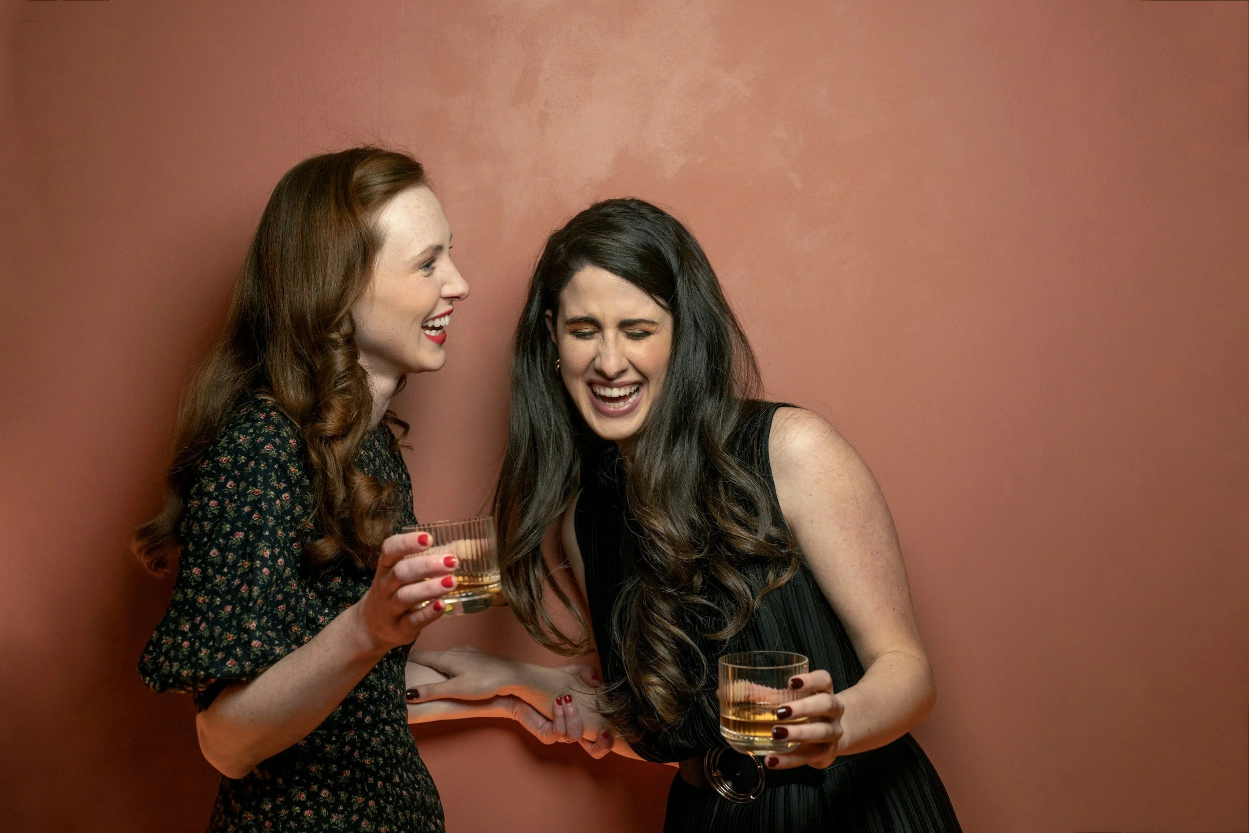 Two women at a party laughing and holding glasses of whiskey against a pink wall.