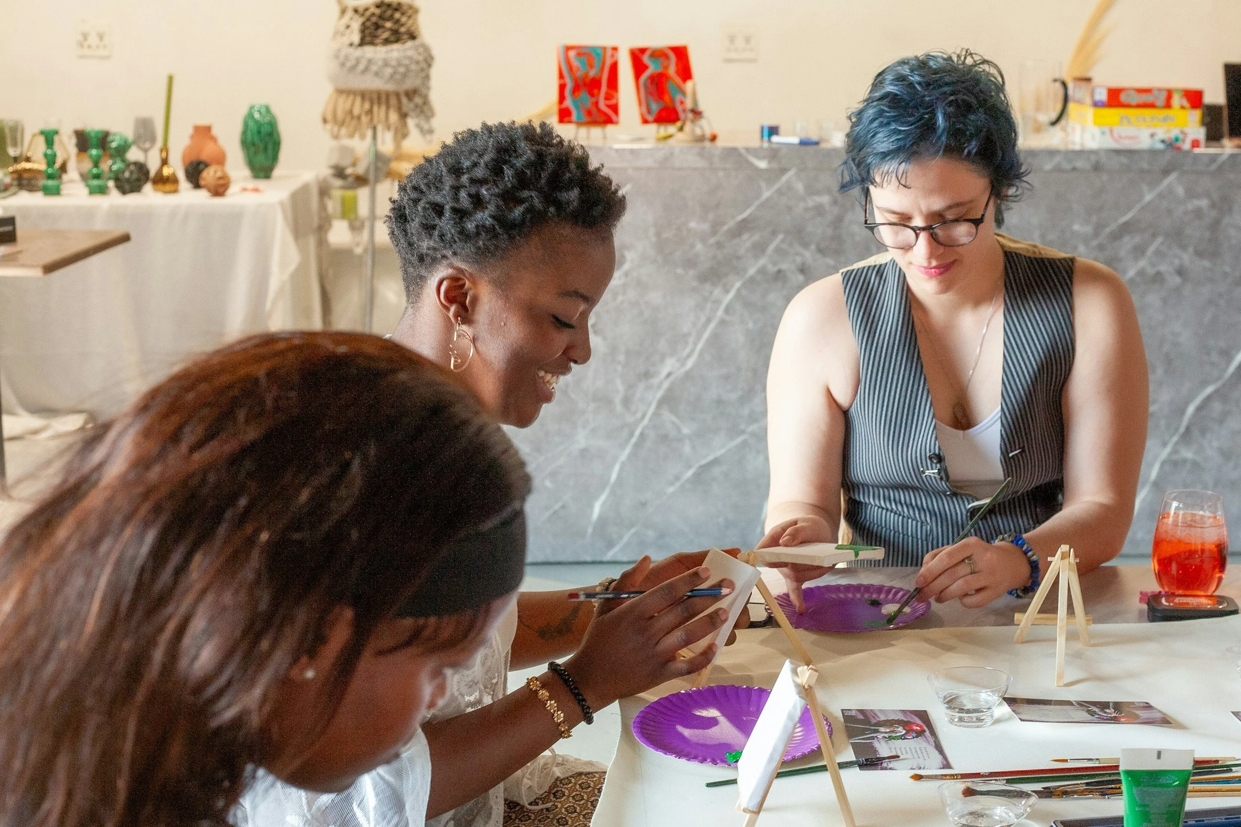 Three women seated at a table, engaging with art or craft activities. In the background, shelves display vases, artwork, and books. The women are focused, smiling, and using various tools and materials.