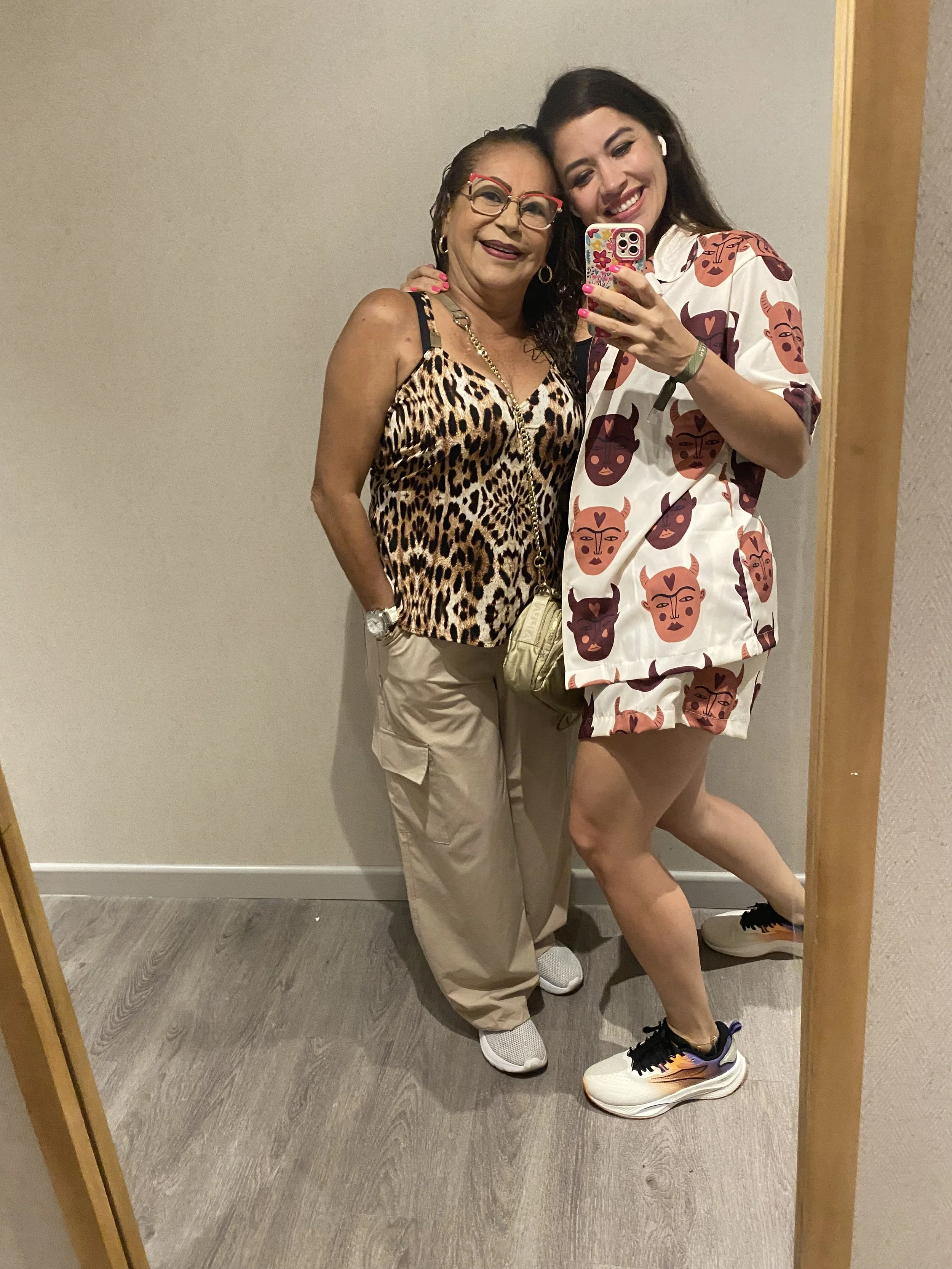 Two women taking a selfie in front of a mirror, smiling and embracing each other. The woman on the left wears a leopard-print top, beige pants, and glasses. The woman on the right wears a matching shirt and shorts with a face and horn pattern, and athletic shoes.