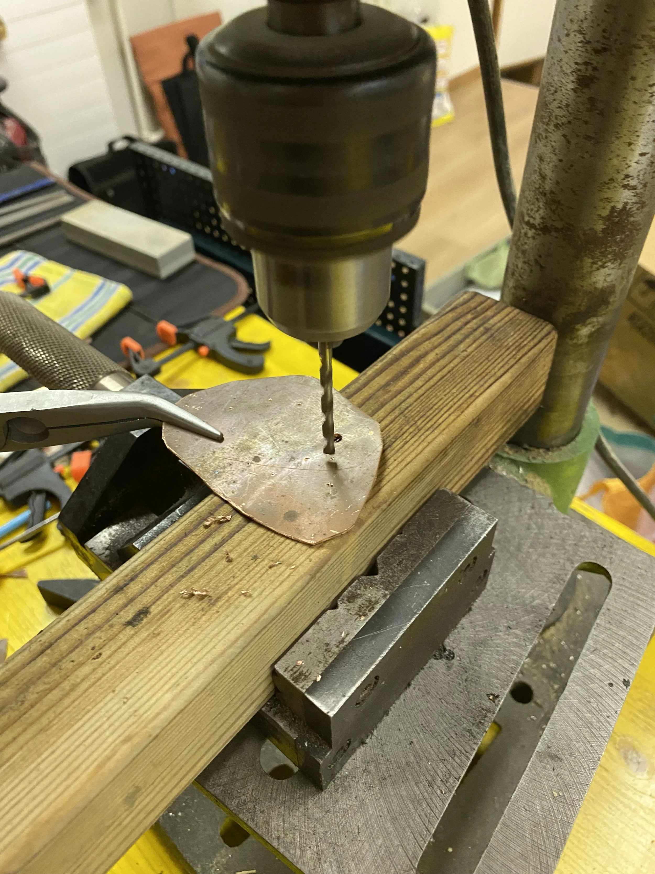 Close-up of a drill press machine with a drill bit, working on a metal piece secured on the worktable. The background shows workshop tools and equipment.