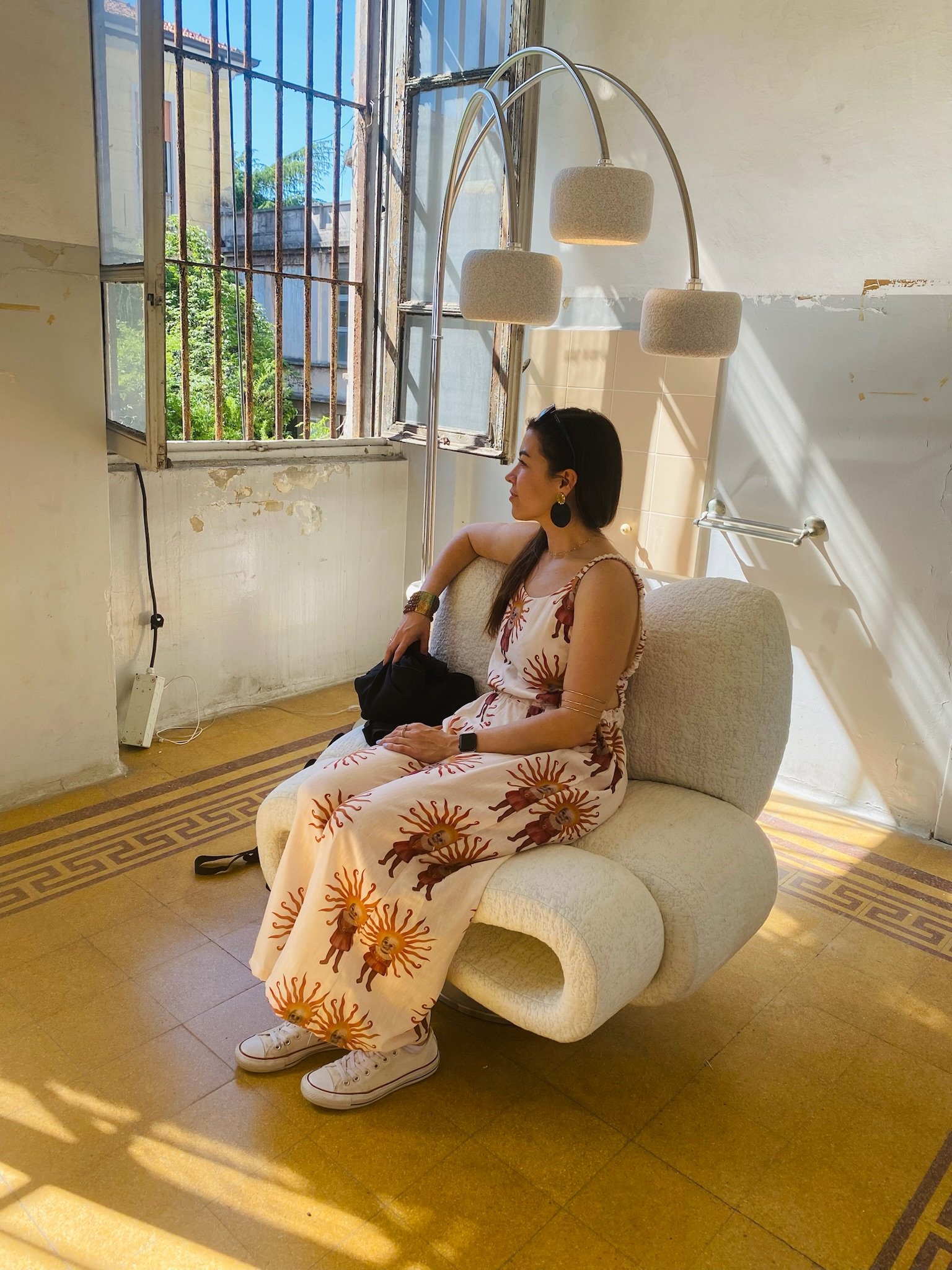 A woman sitting on a beige, plush armchair near a window with peeling paint on the walls. She is wearing a sundress with a lion sun pattern, sneakers, and large earrings, and is looking to the side with a relaxed expression. Sunlight streams through the open window, casting shadows on the wall.