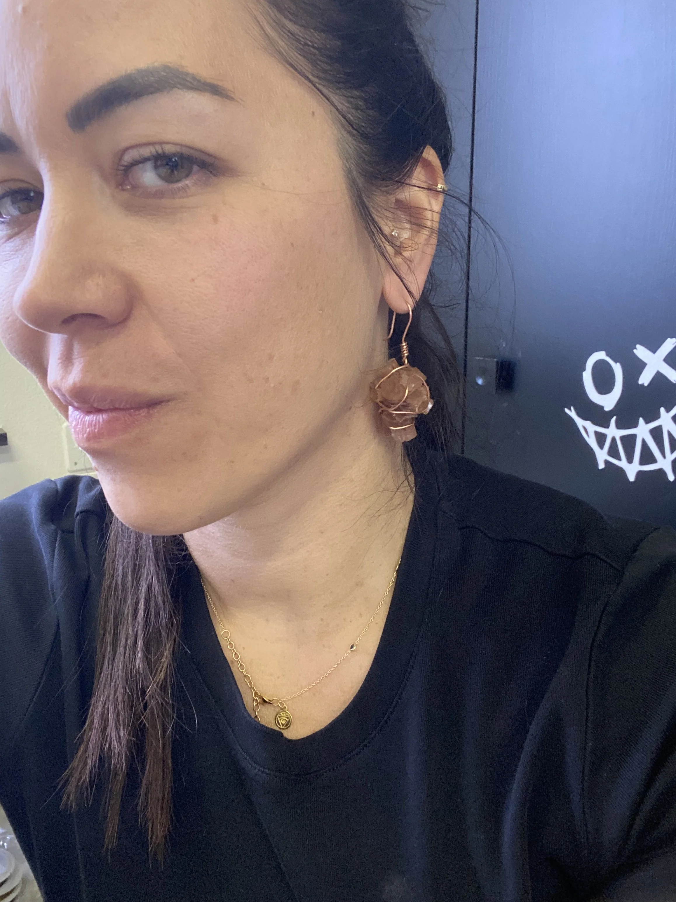 Close-up of a woman wearing gold jewelry, including a necklace with a shield-shaped pendant and earrings with a large amber-colored stone. She has long dark hair, painted eyebrows, light eye makeup, and a neutral facial expression. The background includes a black surface with a white skull design.