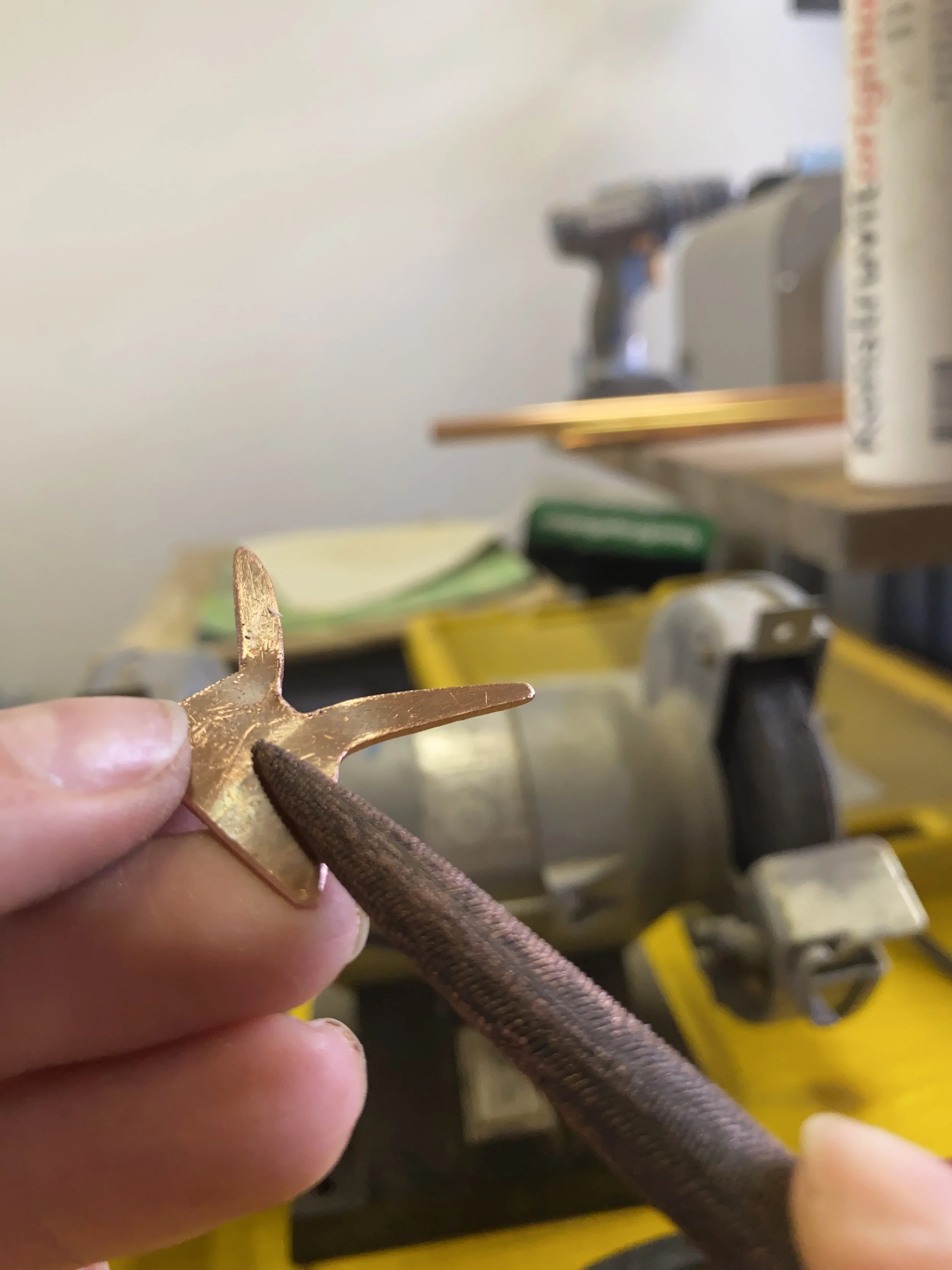 Close-up of a person's hand holding a brass anchor-shaped object with holes, with a partially visible brown textured tool in the background.