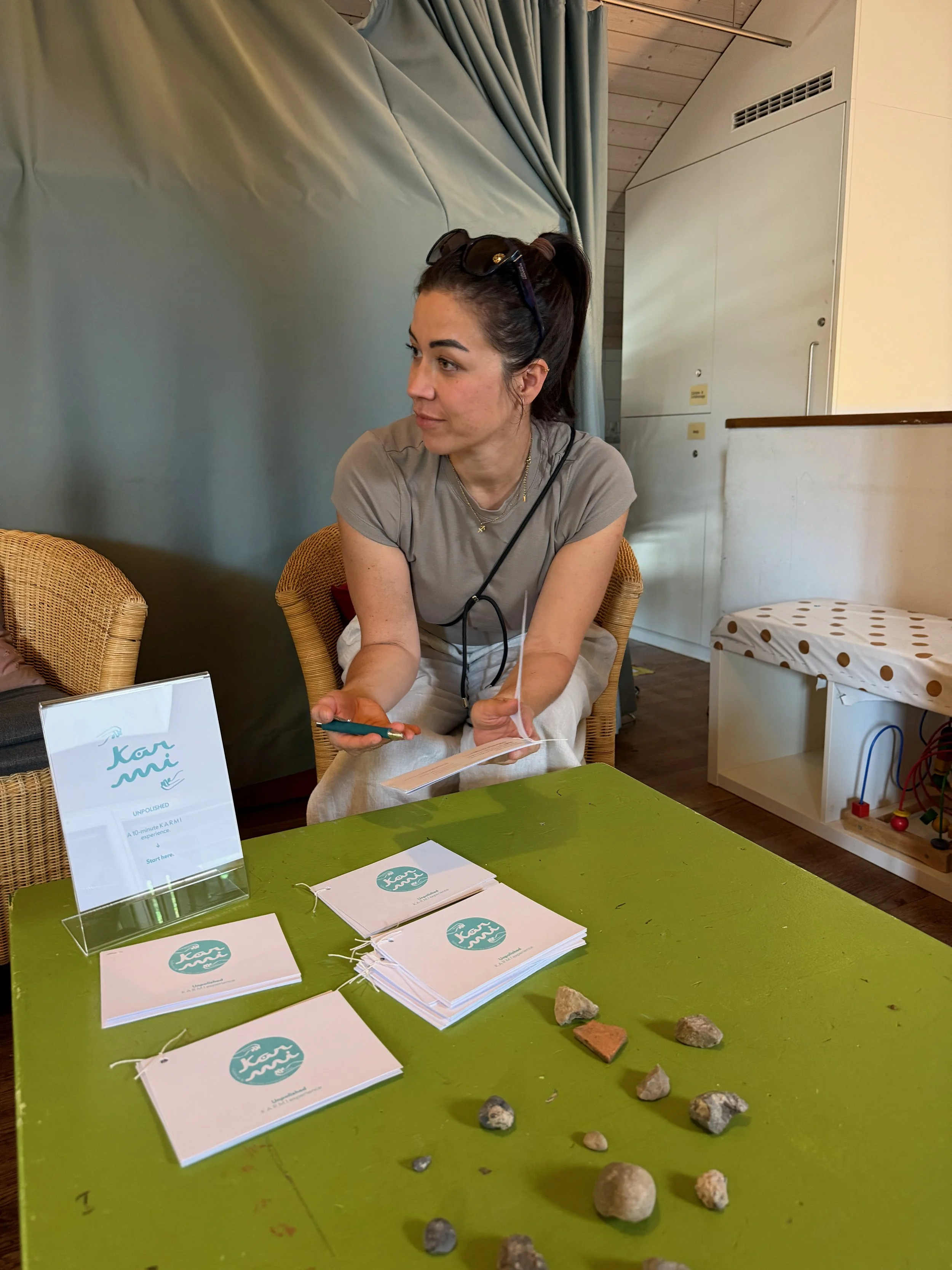 A woman with dark hair tied back and sunglasses on her head is sitting at a green table with brochures, rocks, and rocks on the surface. She is holding a booklet and looking to her left. The background features a corner with a curtain, a white wall with vents, and a play area with beads and wires.