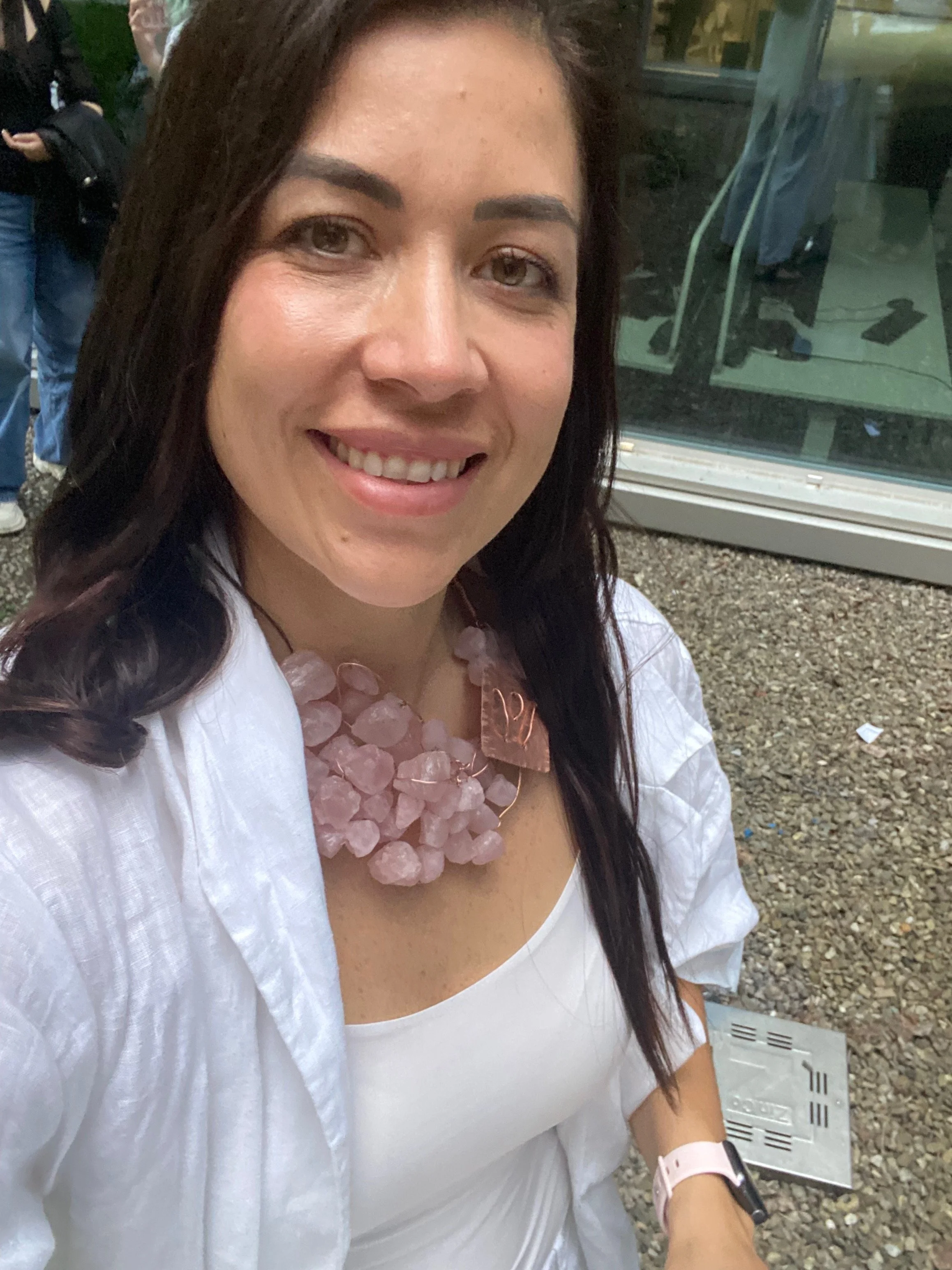 A woman smiling at the camera is wearing a large pink gemstone necklace and a white off-shoulder top. She has long dark hair and is standing outside near a glass building.