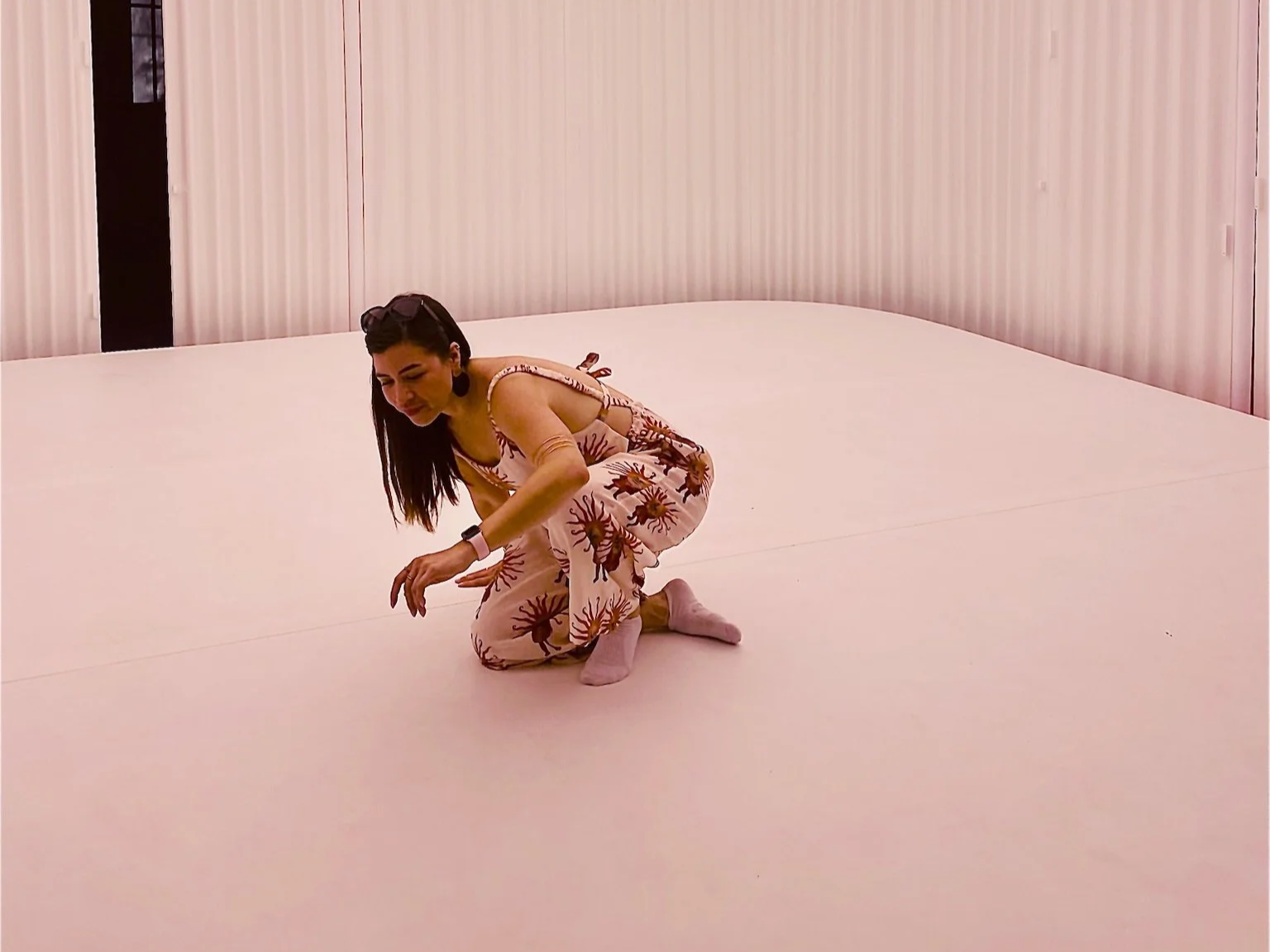 Woman crouching on a large pink floor with pink walls, wearing a patterned jumpsuit and pink socks, with sunglasses on her head and a smartwatch.