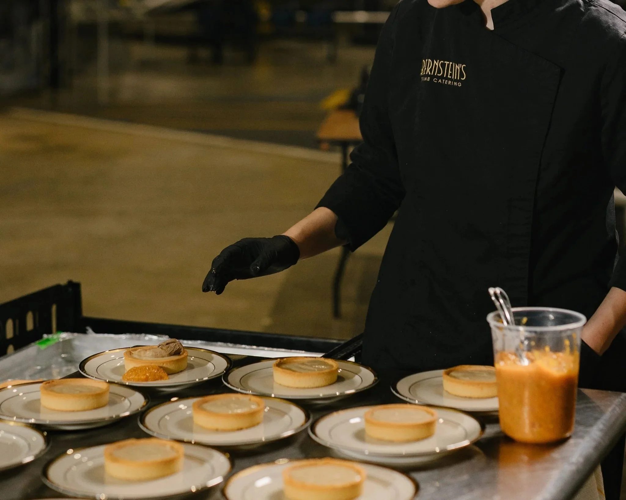 A chef wearing a black uniform and gloves preparing desserts at a catering event. Several plates with cheesecakes and a glass filled with a layered dessert are on the table.