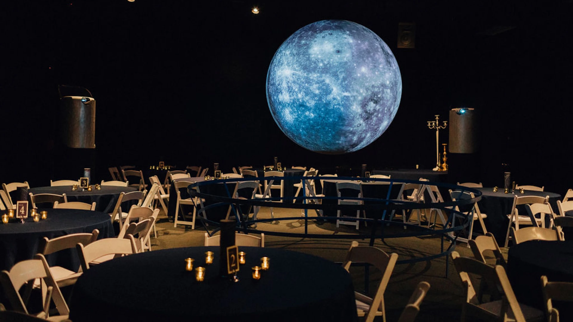 Event space decorated with tables and chairs, large projected image of the moon on a dark wall, and ambient lighting.