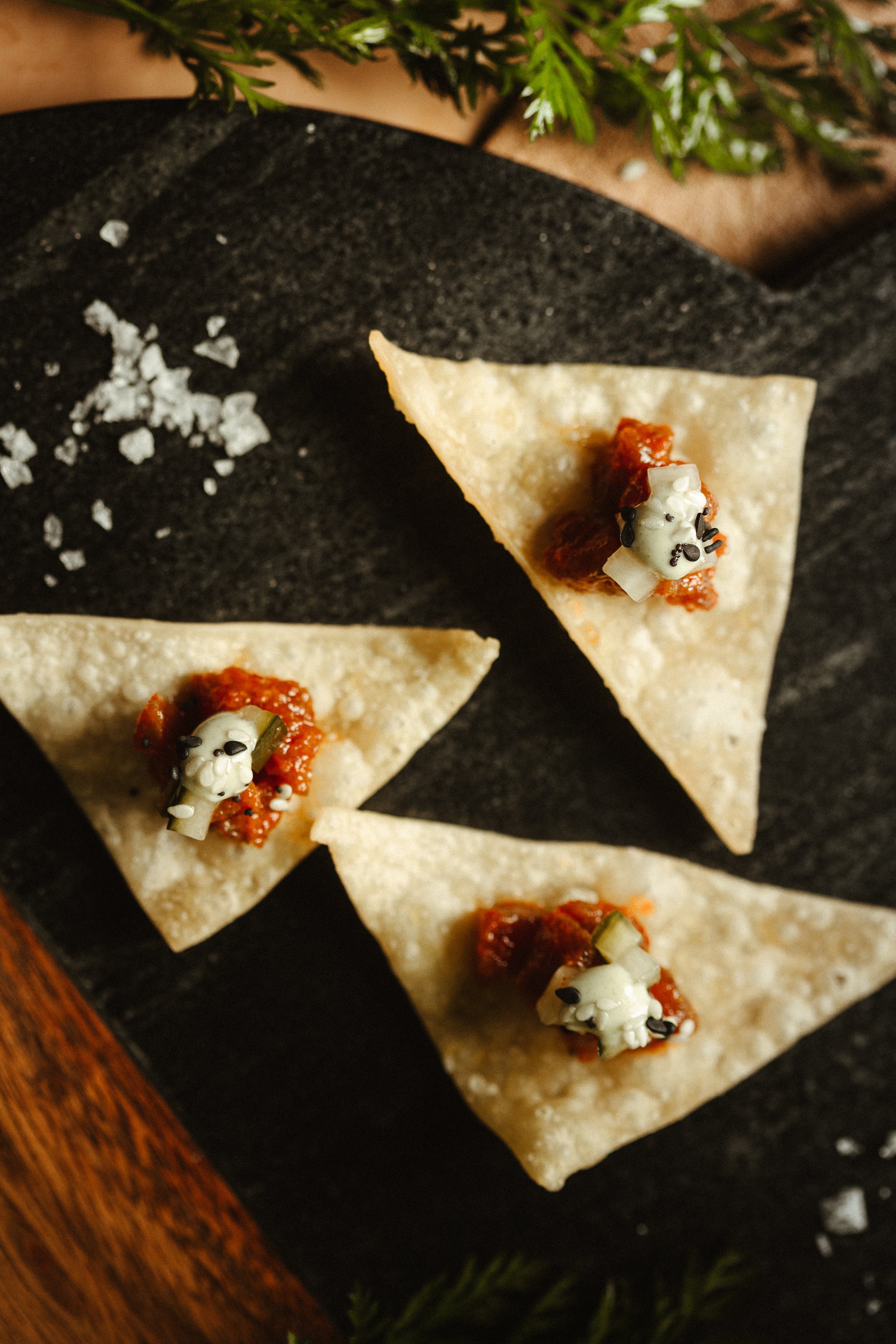 Close-up of tortilla chips topped with tomato salsa and dollops of sour cream garnished with black sesame seeds, set on a black slate serving platter with sprigs of greenery in the background.
