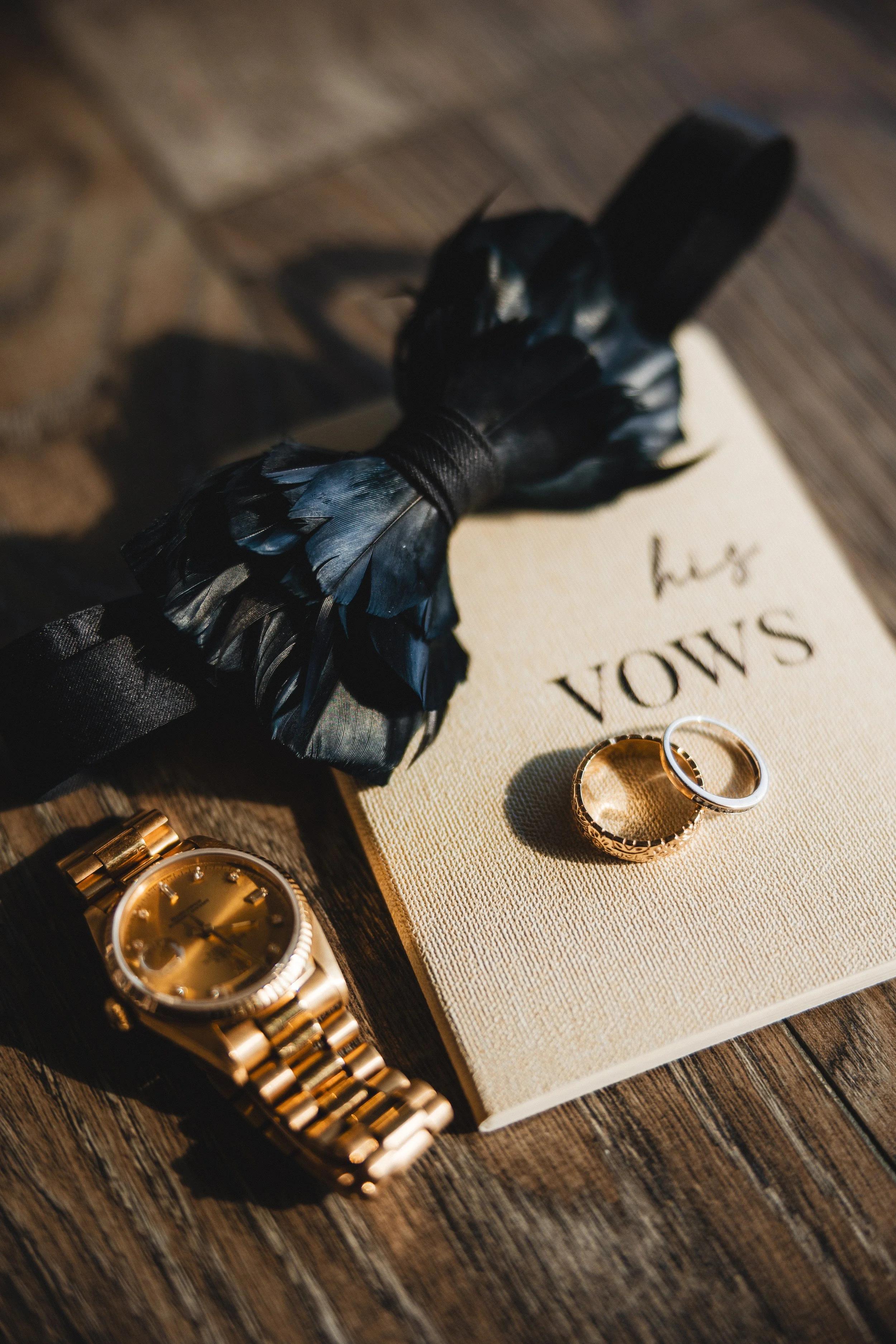 A close-up of gold jewelry including a wristwatch and rings on a wooden surface, with a book titled 'Vows' and a black feathered object nearby.