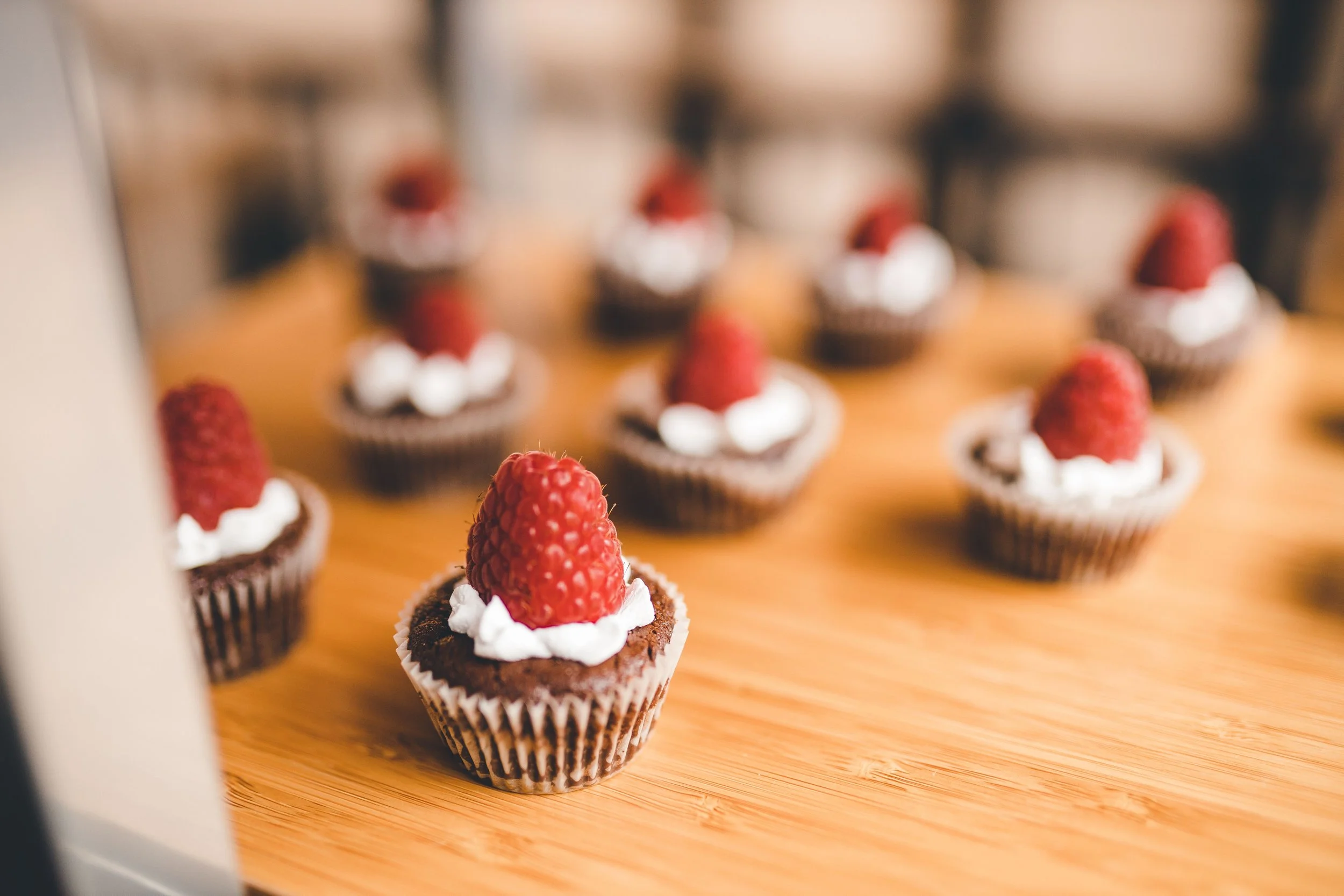 Close-up of a cupcake with chocolate frosting, whipped cream, and a raspberry on top, on a wooden surface with additional cupcakes blurred in the background.