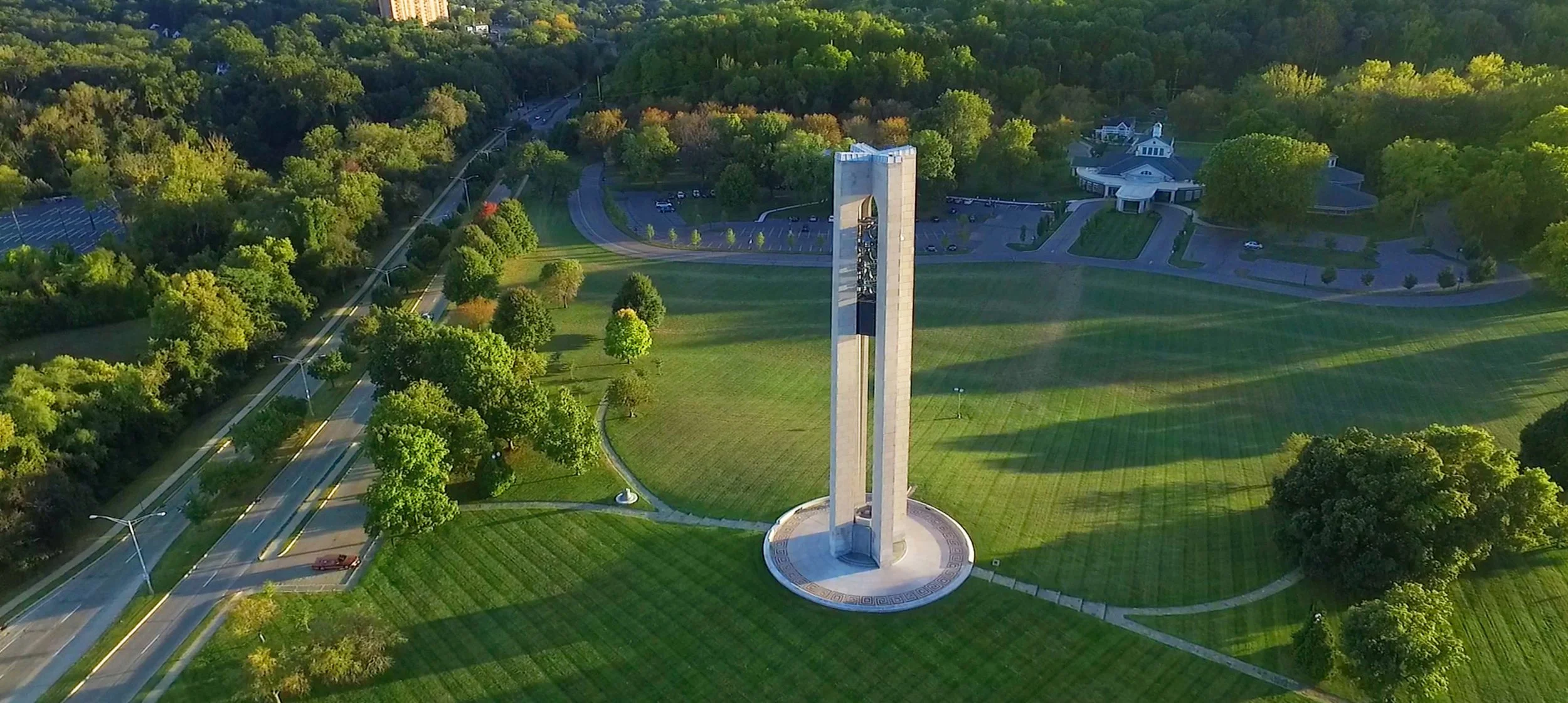 Aerial view of a park with a tall monument at the center, surrounded by green grass, trees, parking lots, and nearby buildings in the background.