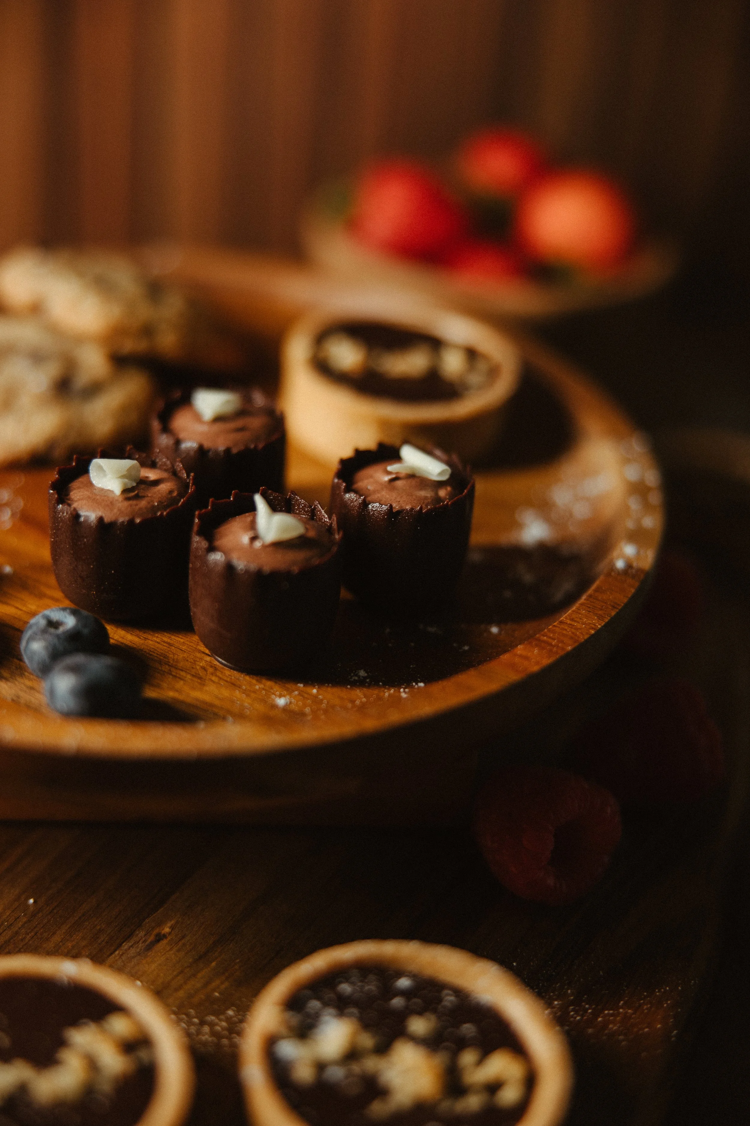 Variety of small, round desserts topped with white chocolate shavings on a wooden platter, with berries in the background.