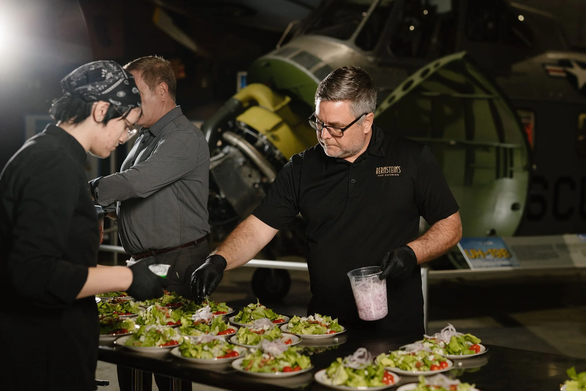 Three people preparing salads on a table in front of a helicopter inside a hangar.