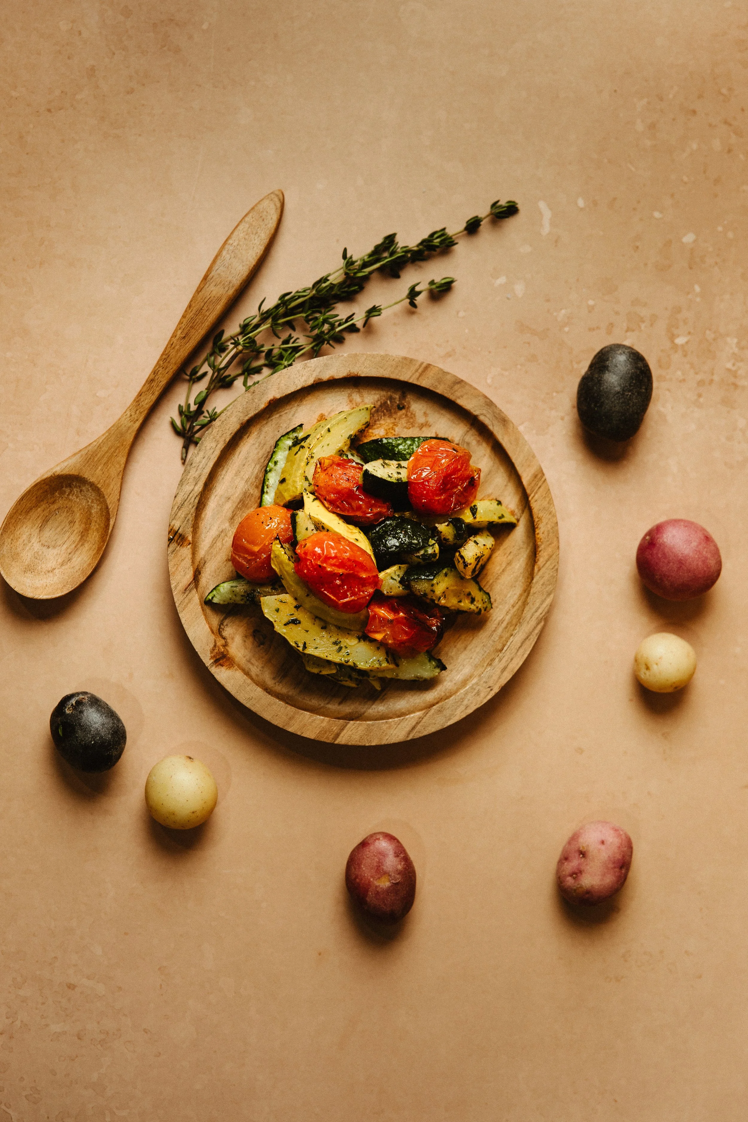 A wooden bowl with roasted cherry tomatoes, sliced zucchini, and herbs, placed on a beige background. Around the bowl, there are various small potatoes and a wooden spoon and a sprig of fresh thyme.