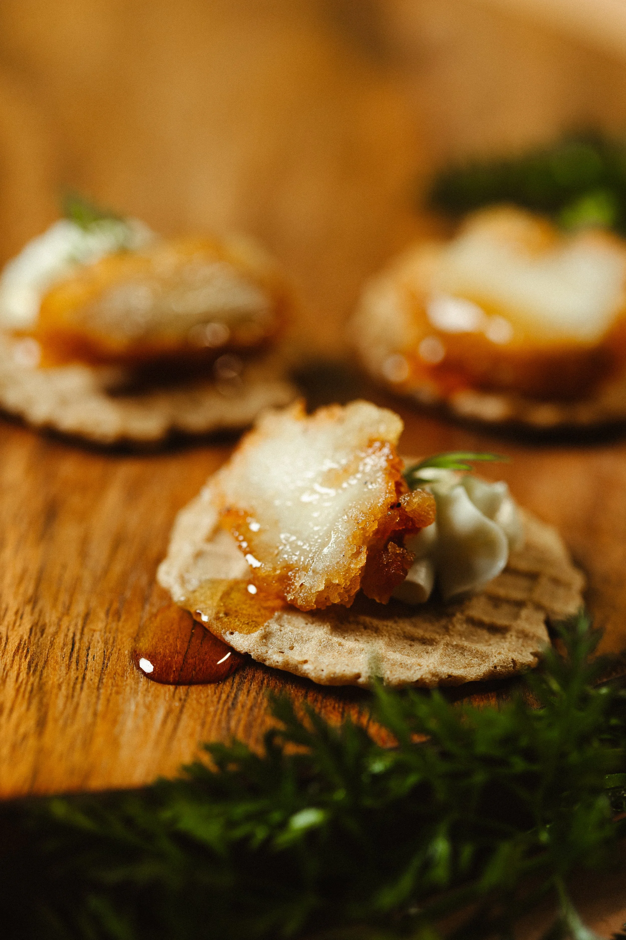 Close-up of a piece of fried chicken on a small round cracker, garnished with fresh herbs, set on a wooden surface.