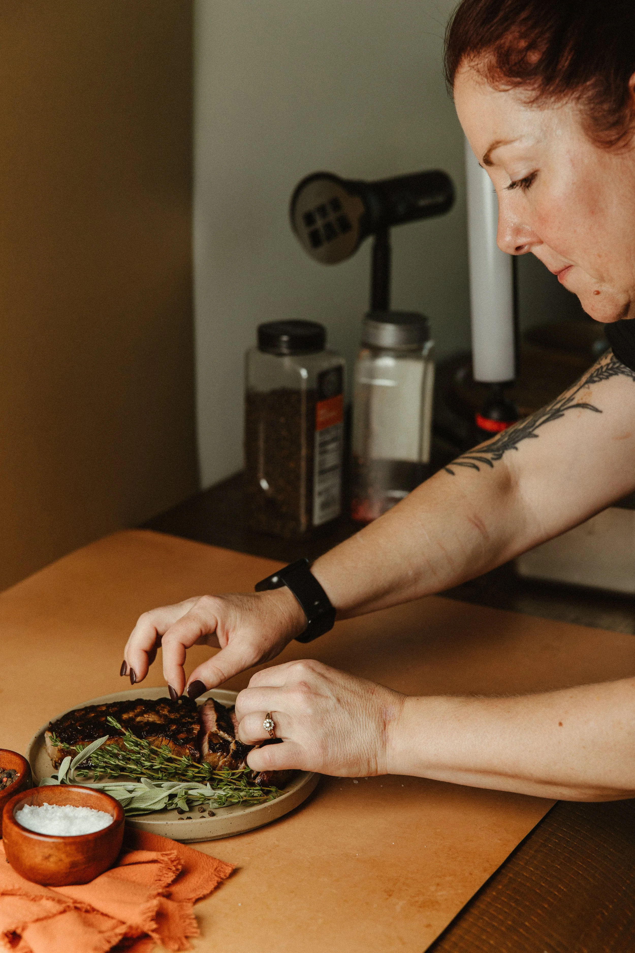 A woman arranging food on a plate with herbs, with spices in jars and a grill in the background.