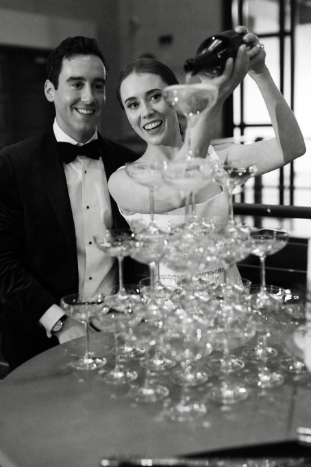 A woman is pouring champagne into a pyramid of glasses while a man watches and smiles during a celebration or event.