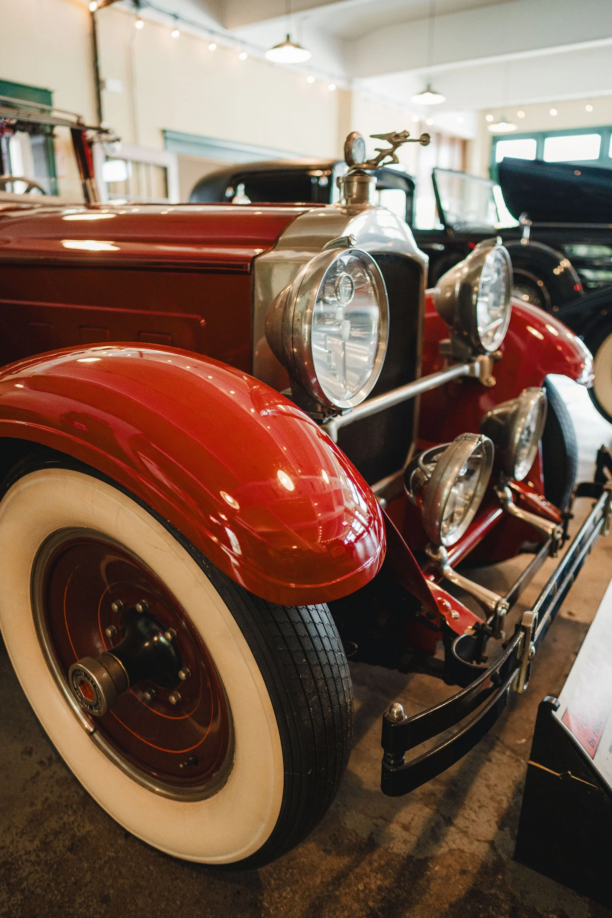 Close-up of a vintage red car with large white tires and chrome details, displayed indoors with other classic vehicles in the background, in a museum or showroom setting.